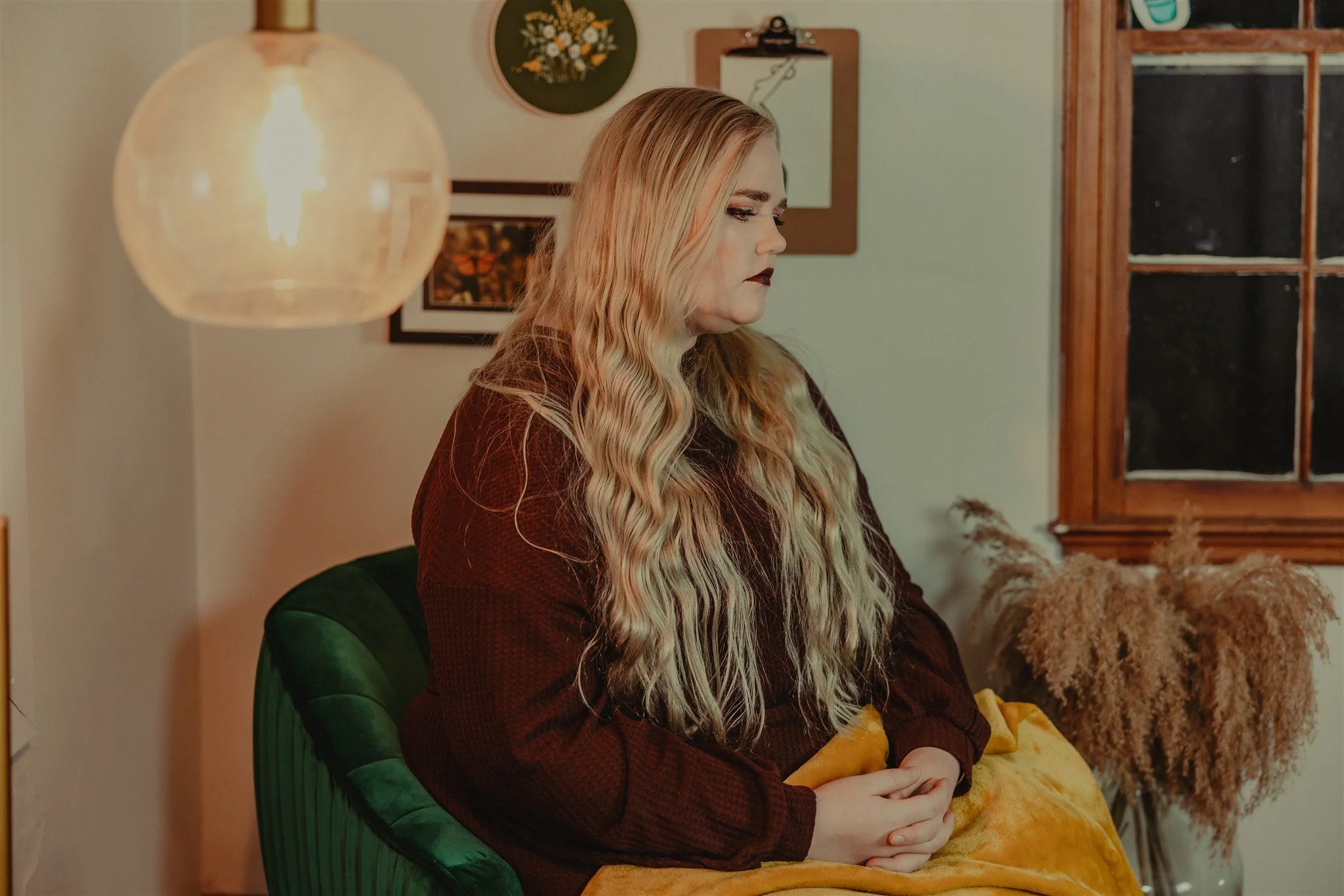 A woman with long, wavy blonde hair and dark lipstick sitting on a green velvet chair in a cozy, decorated room. The room has framed photos and artwork on the wall, a large hanging light during a trauma informed photography session in Columbia, MO.