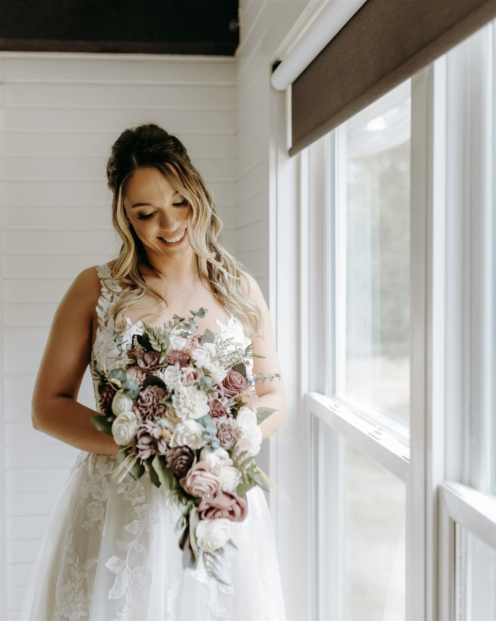 Portrait of a bride in a white lace wedding dress holding a large bouquet of pink, white, and lavender roses, standing near a window at Rolla, Missouri rustic wedding venue, The Village. Photos by Columbia, MO Photographer Liv Strange Photography.