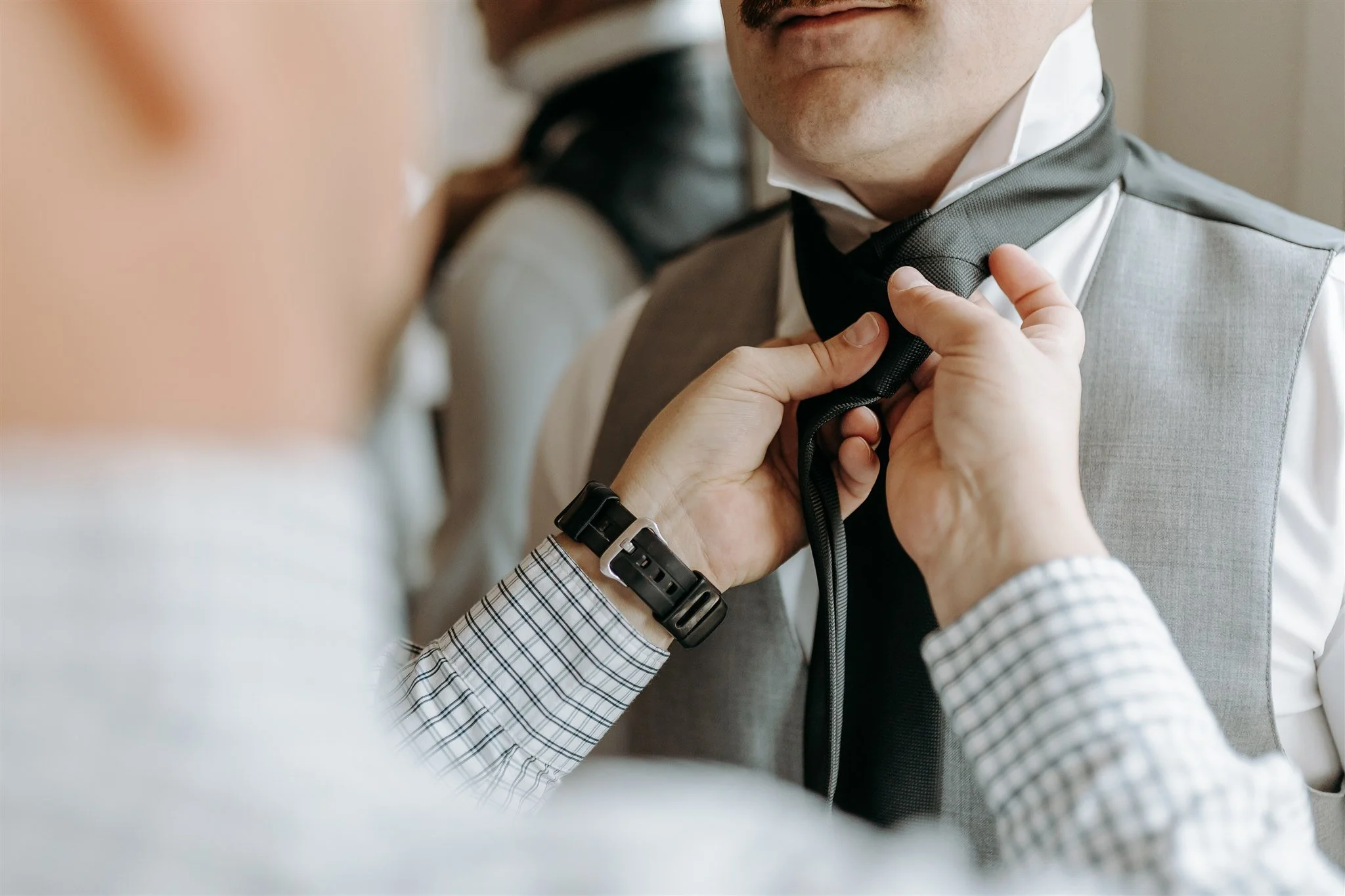 A person adjusting a black tie on a groom wearing a gray vest and white shirt, with another person nearby at Rolla, Missouri rustic wedding venue, The Village. Photos by Columbia, MO Photographer Liv Strange Photography.