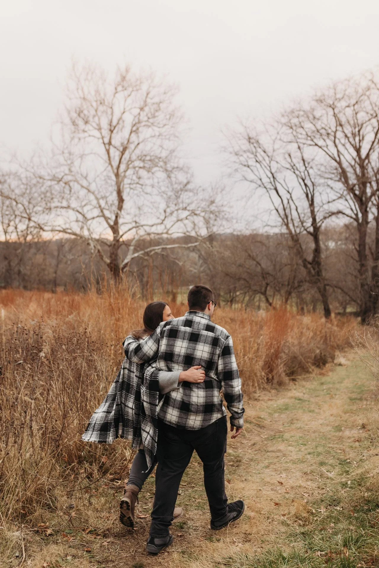 A couple walking hand in hand on a dirt path through a field of dry grass and trees in autumn or winter, dressed in plaid shirts and jackets at their engagement photography session at Queeny Park in STL.