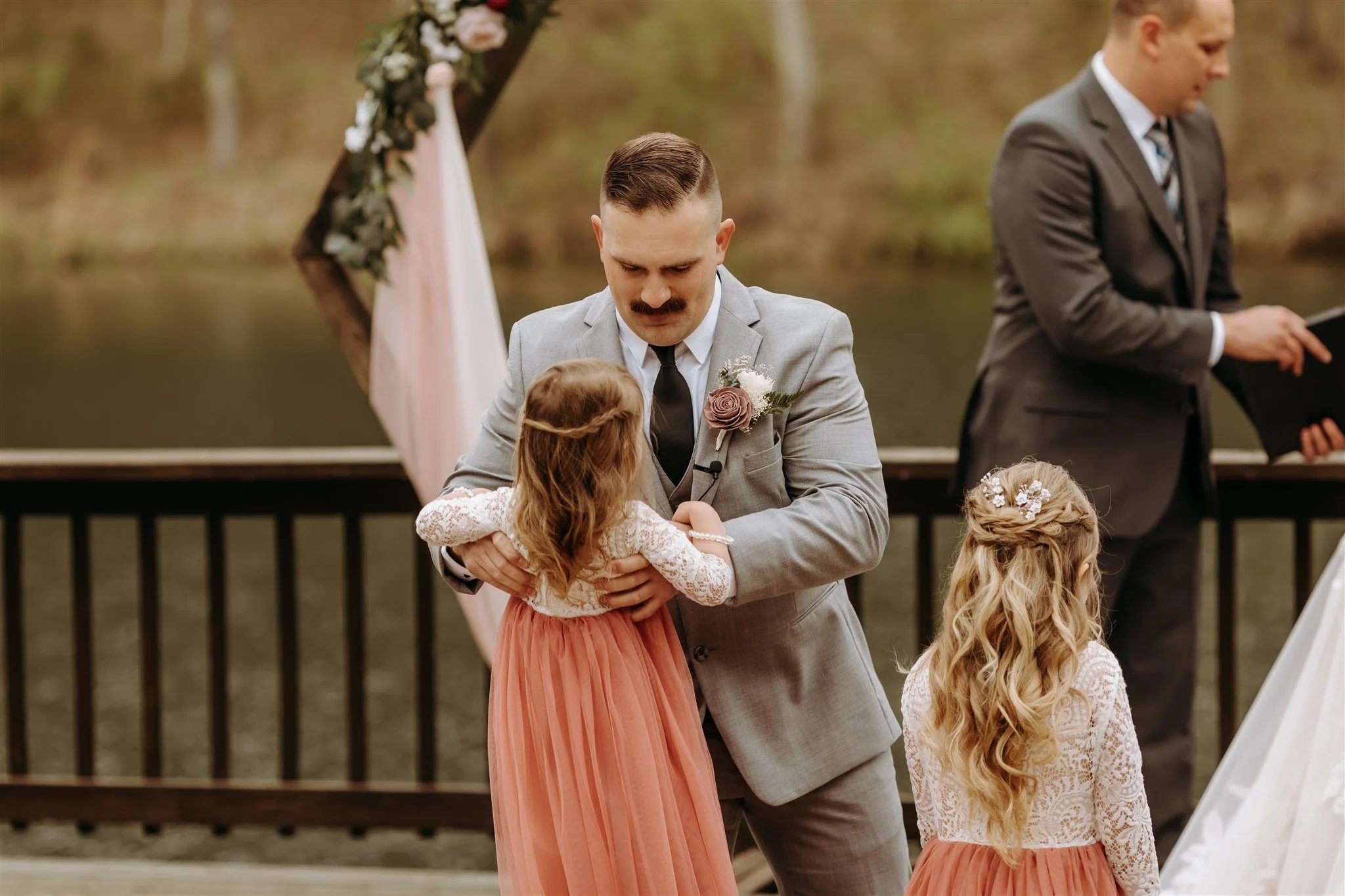 A man in a gray suit, holding a young girl in a long pink dress, standing outdoors near water during a wedding ceremony at Rolla, Missouri rustic wedding venue, The Village. Photos by Columbia, MO Photographer Liv Strange Photography.