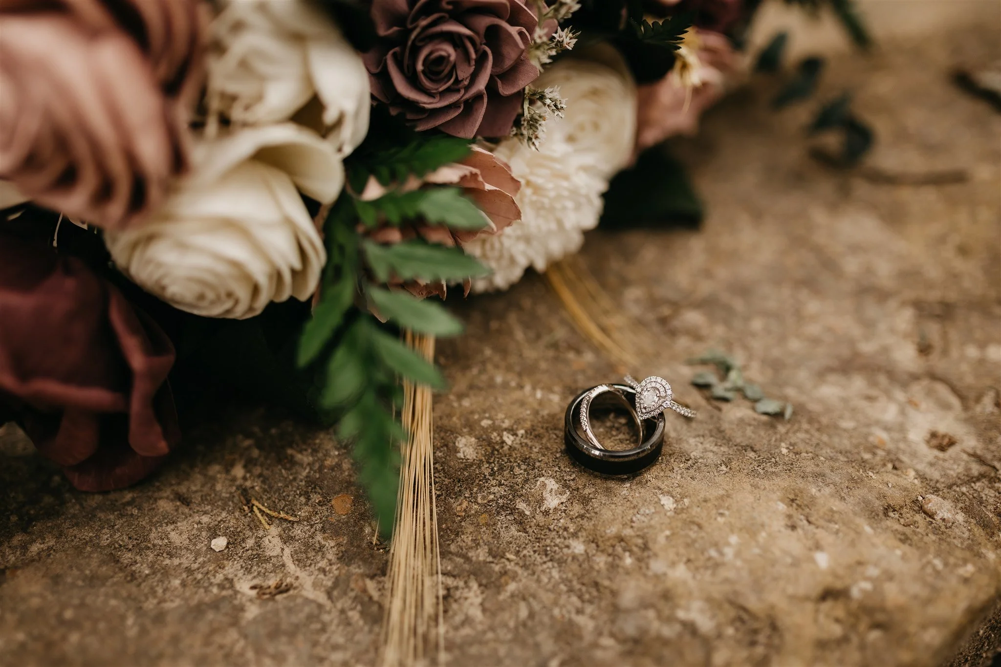 A close-up of wedding rings placed on the ground next to a bouquet of flowers, consisting of white and purple roses at Rolla, Missouri rustic wedding venue, The Village. Photos by Columbia, MO Photographer Liv Strange Photography.