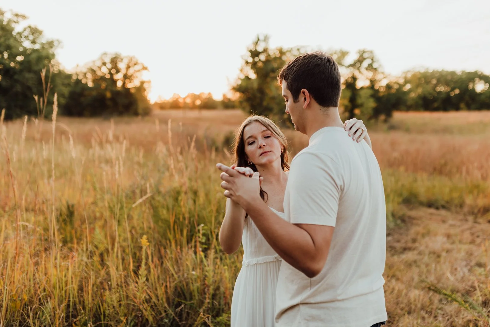 A couple is dancing on a field at sunset, holding hands and gazing at each other, with tall grass and trees in the background during their couples photography session at Rock Bridge State Park in Columbia, MO.