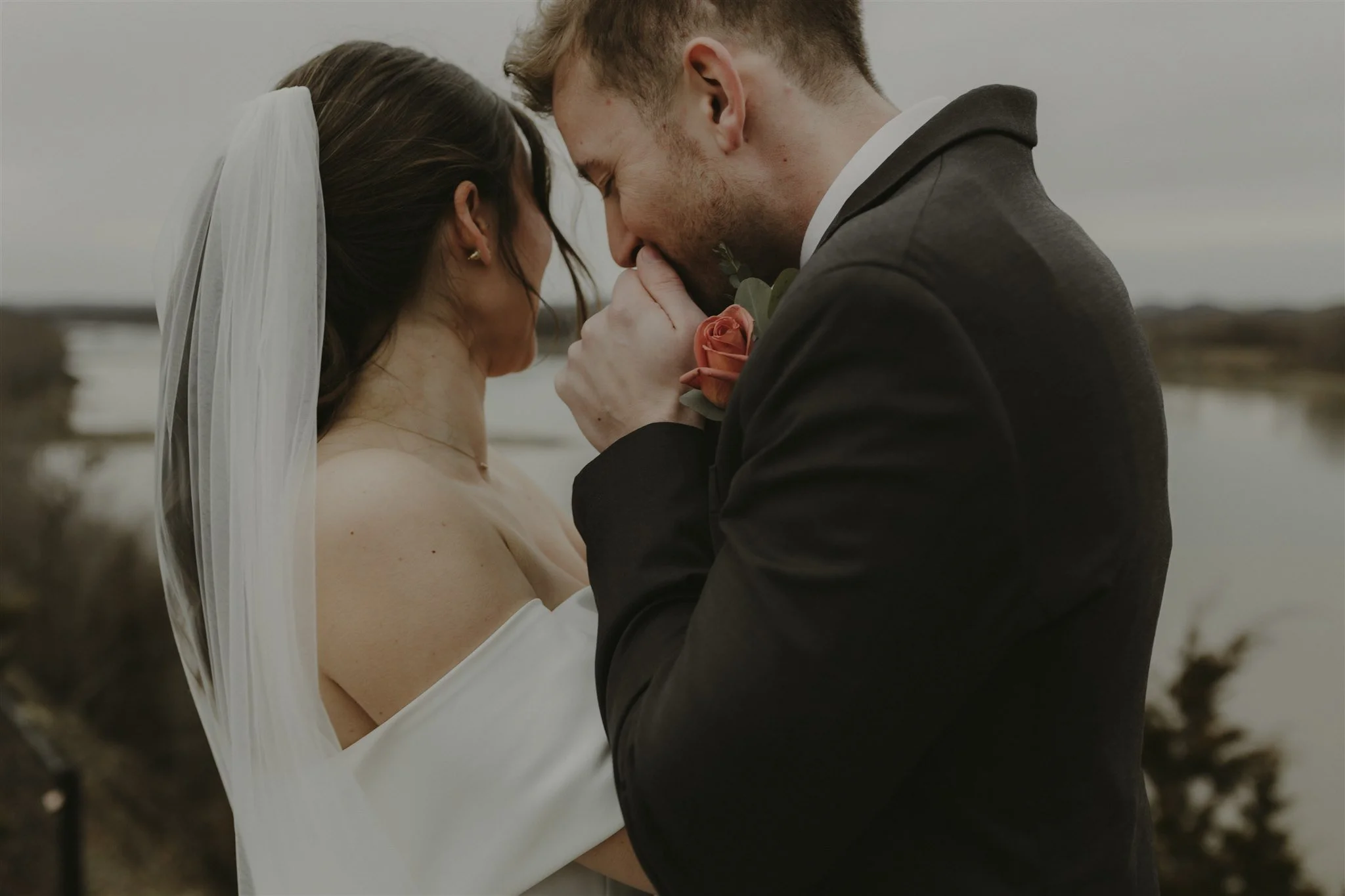A bride and groom sharing a tender moment outdoors, with the groom kissing the bride's hand as she leans in with her eyes closed, a river in the background, and overcast weather at wedding venue in Hermann, MO, Hermann Hills.