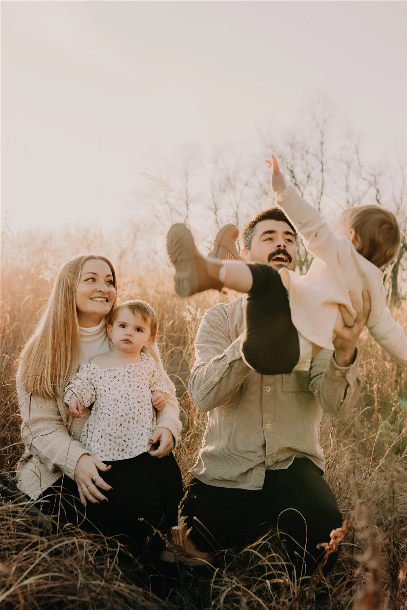 A family of four enjoying time outdoors in a field during sunset. The father is lifting a young girl in the air, and the mother is sitting with her daughter on her lap, during their Columbia, MO family photography session at Rock Bridge State Park.