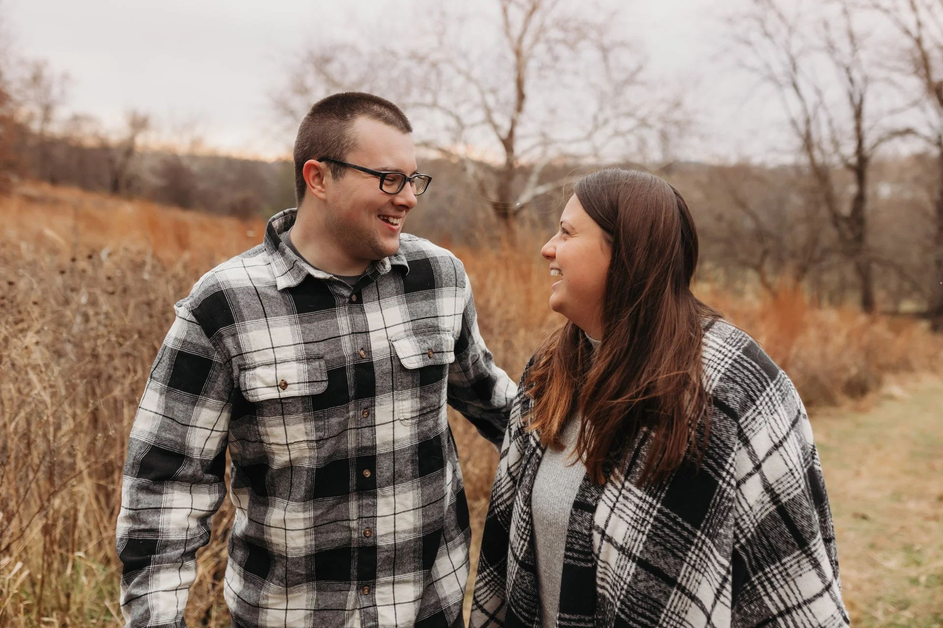 A man and woman standing outdoors in a field of tall, brown grass, smiling and looking at each other on a cloudy day with leafless trees in the background at their engagement photography session at Queeny Park in STL.