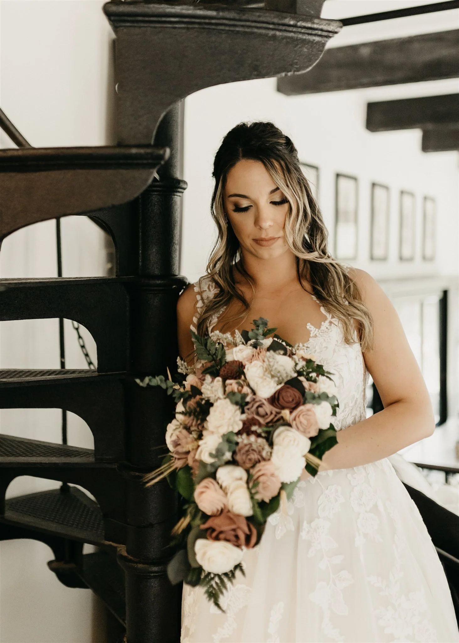 Portrait of a bride in a white lace wedding dress holding a large bouquet , standing near a black staircase inside a building at Rolla, Missouri rustic wedding venue, The Village. Photos by Columbia, MO Photographer Liv Strange Photography.