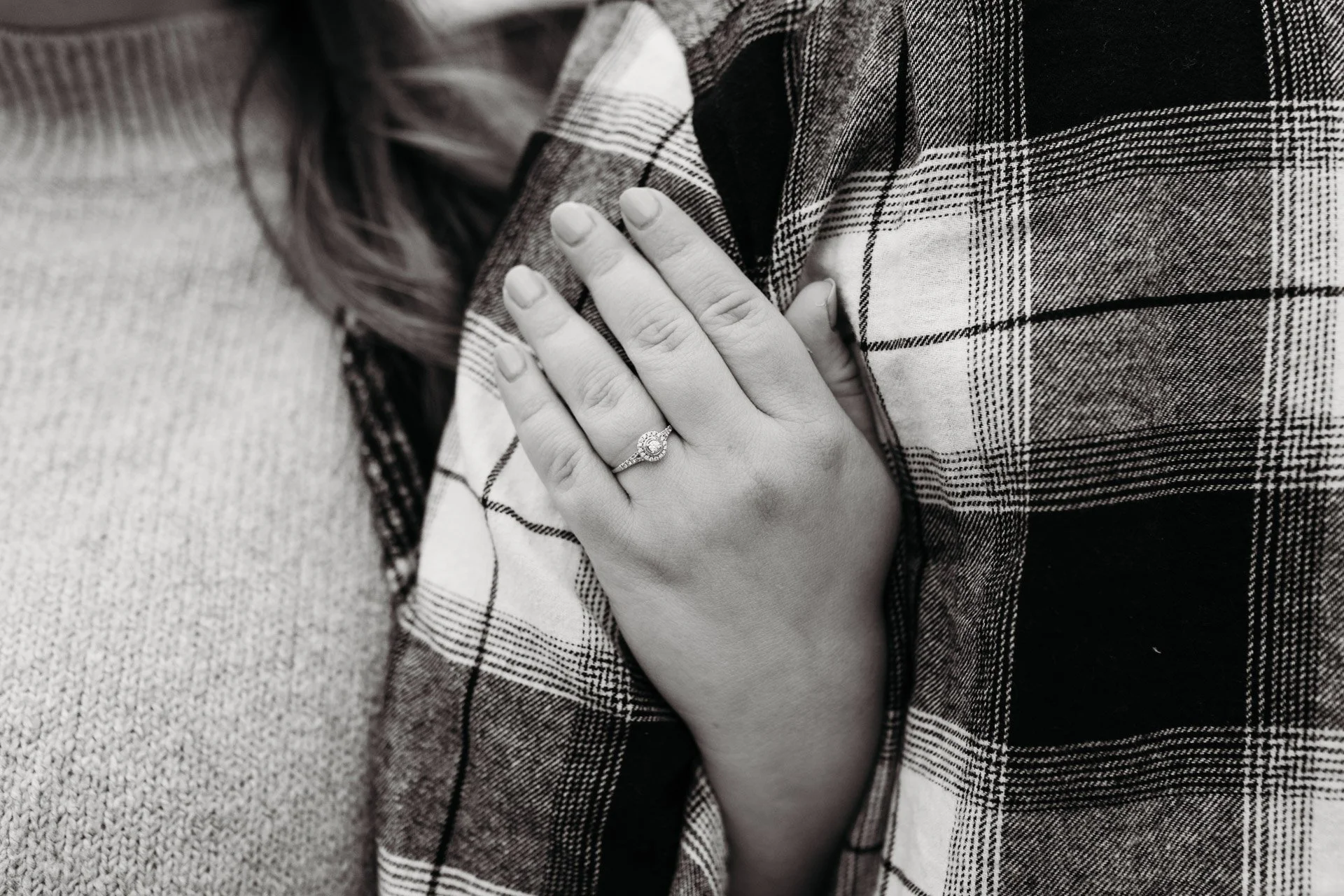 Close-up of a woman's hand with a wedding ring resting on her chest, with a plaid shirt layered over a sweater at their engagement photography session at Queeny Park in STL.