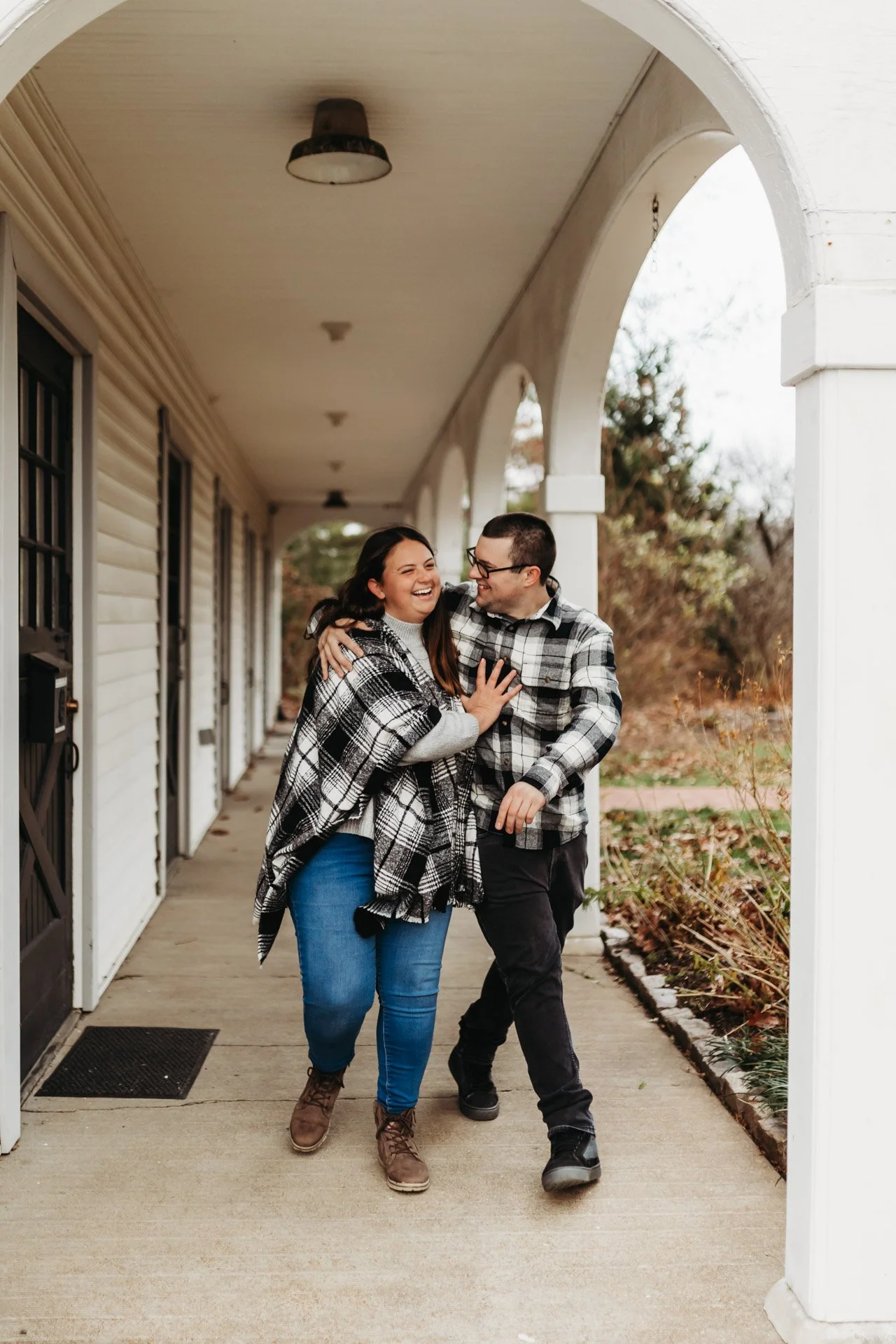 A happy couple walking arm in arm along a porch, smiling and laughing together, dressed in black and white plaid shirts during fall at their engagement photography session at Queeny Park in St. Louis, MO