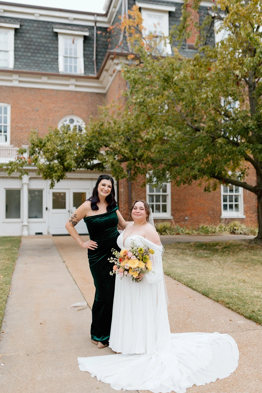 Two women, one in a green velvet dress and the other in a white wedding gown, standing outside in front of a brick building at an LGBTQ+ wedding in Columbia, MO at wedding venue, Stephens College Senior Hall.