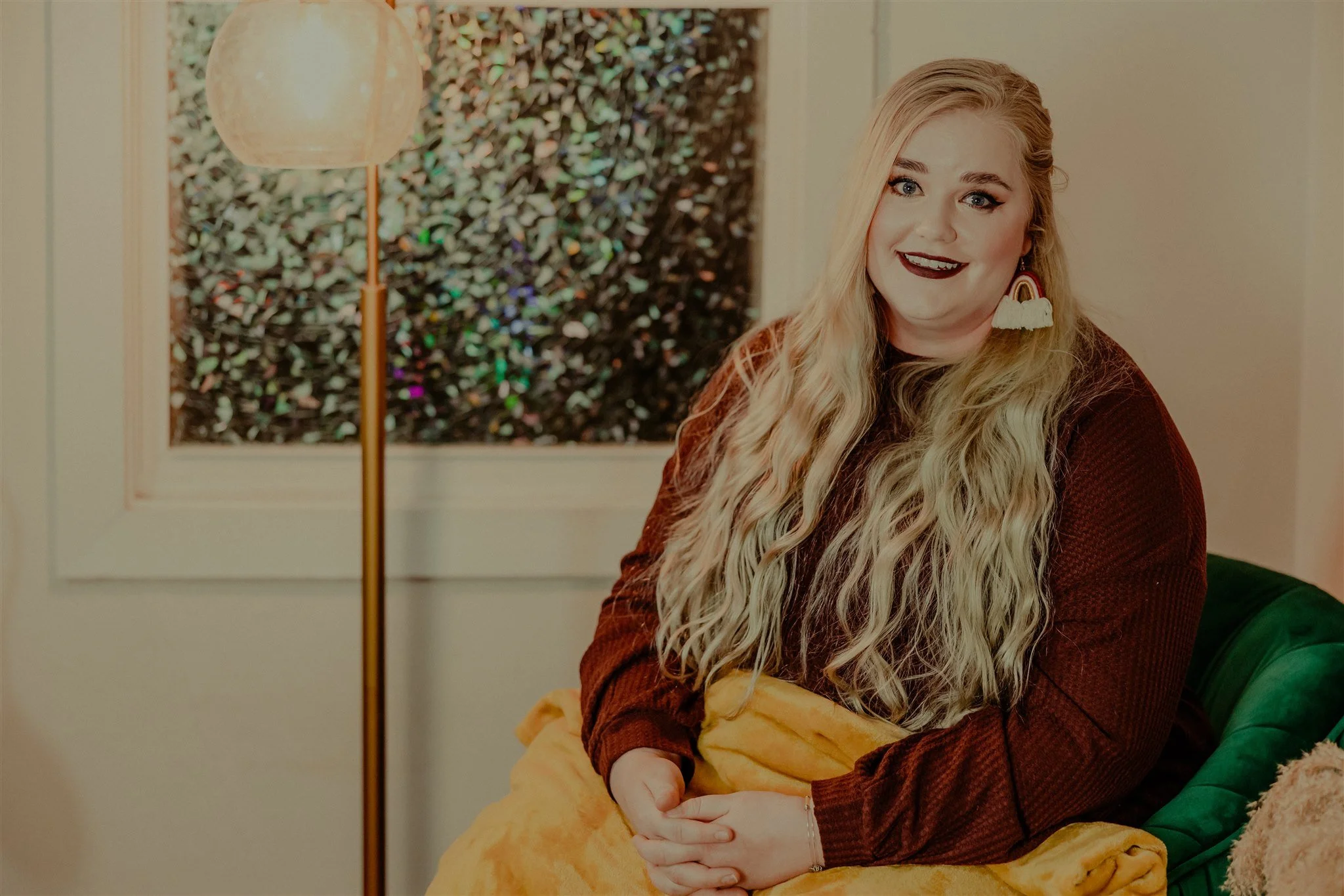 A woman with long blonde curly hair, wearing a brown top and rainbow-shaped earrings, sitting on a green velvet chair with a yellow blanket during a trauma informed photography session in Columbia, Missouri.