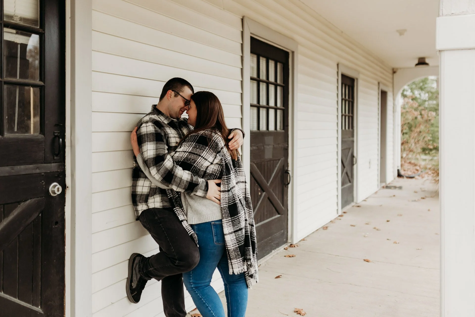 A couple dressed in plaid shirts embrace and smile while leaning against a white wall outside a building with barn-style doors at their engagement photography session at Queeny Park in STL.
