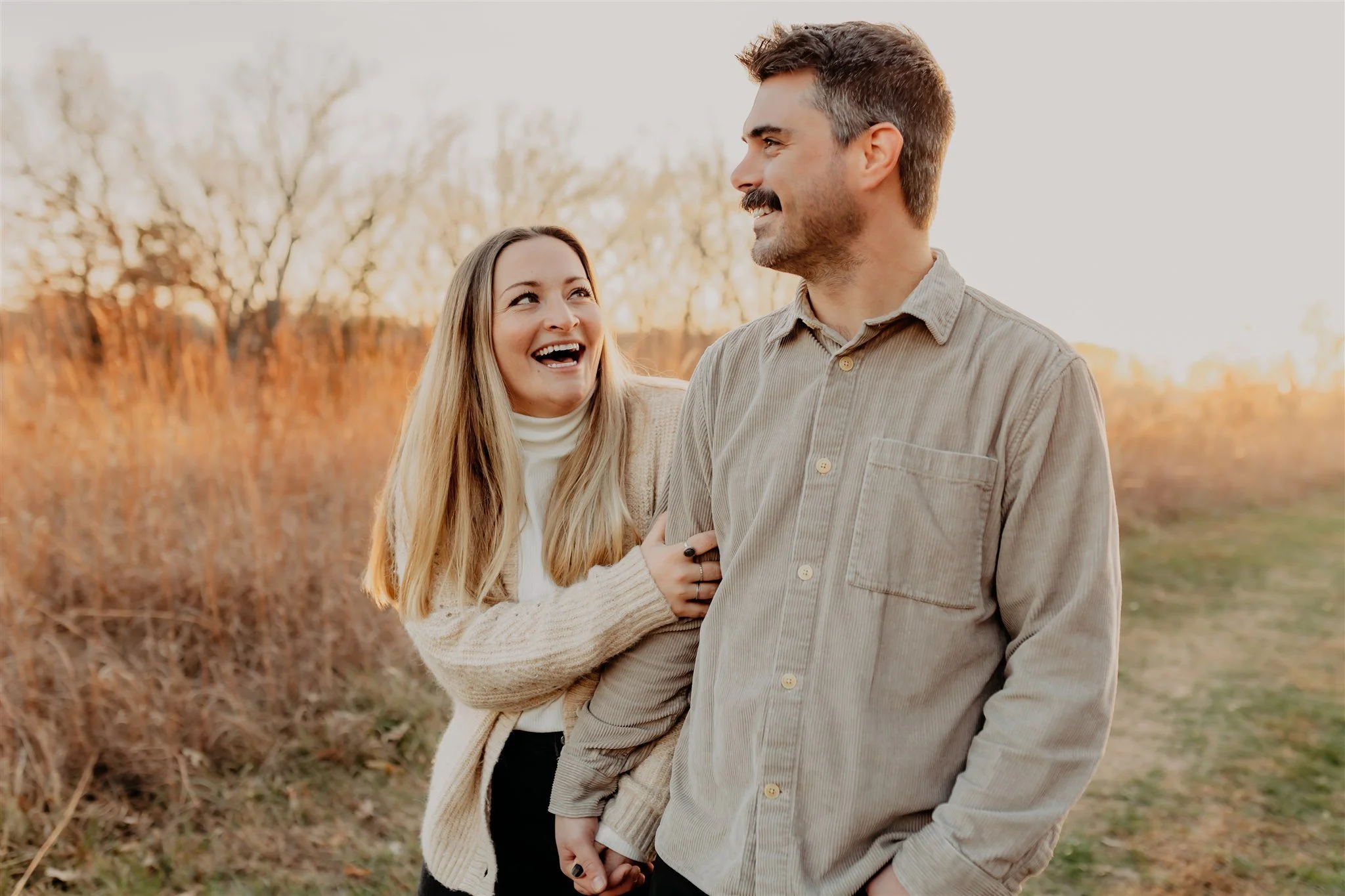A smiling couple walking outdoors in a field during sunset, holding hands with the woman playfully tugging on the man's arm during their Columbia, MO couples photography session at Rock Bridge Memorial State Park.