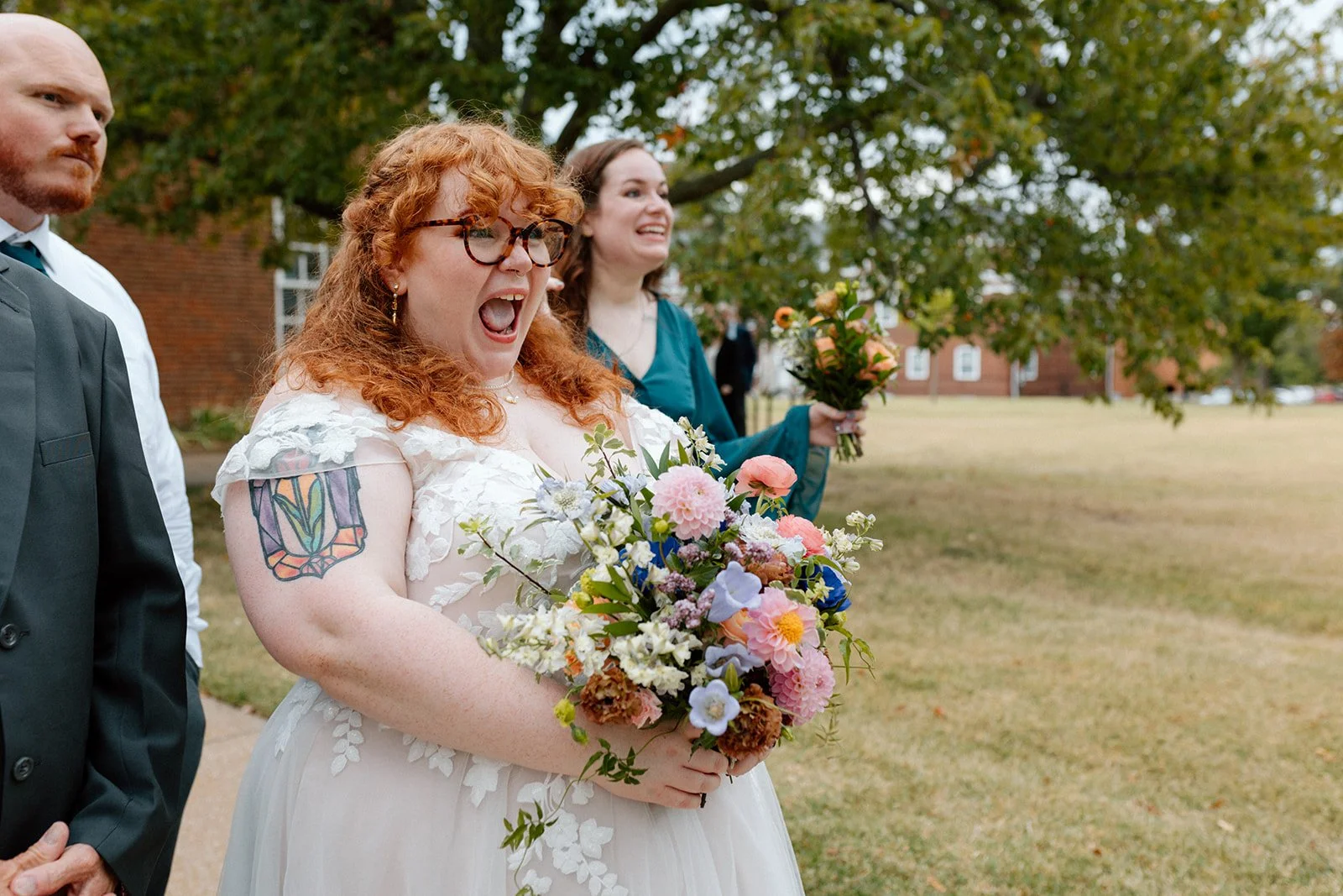 A group of people standing outdoors in a park, with a bride in a wedding dress holding a bouquet of flowers, smiling and celebrating at an LGBTQ+ wedding in Columbia, MO at wedding venue, Stephens College Senior Hall.