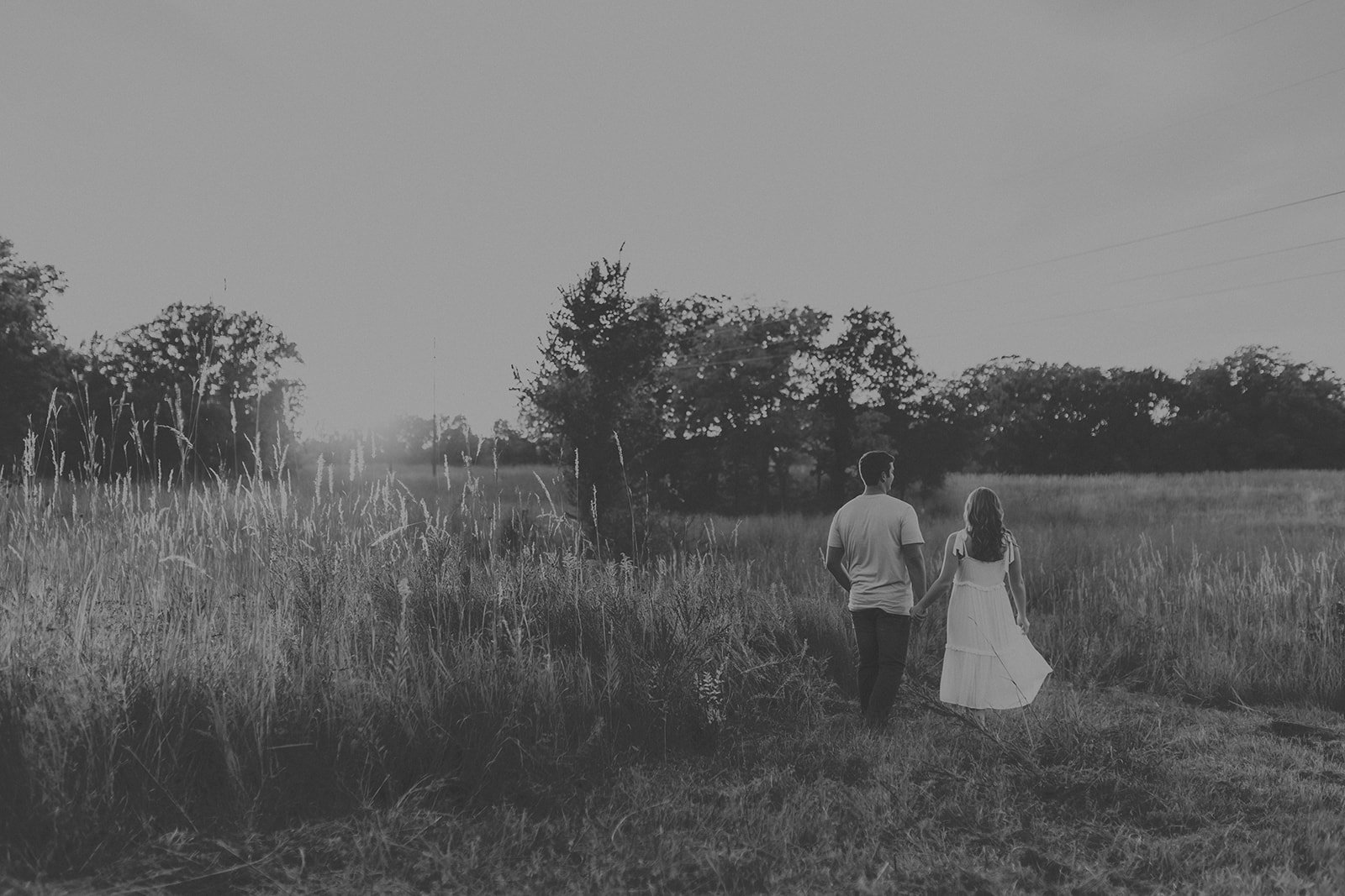 A black and white photo of a couple holding hands walking through a grassy field with trees in the background during sunset during their couples photography session at Rock Bridge State Park in Columbia, MO.