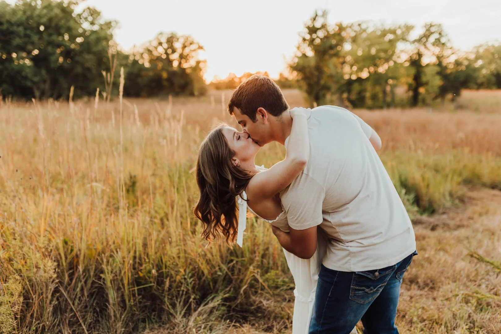 A couple sharing a kiss in a field at sunset, with trees in the background during their couples photography session at Rock Bridge State Park in Columbia, MO.