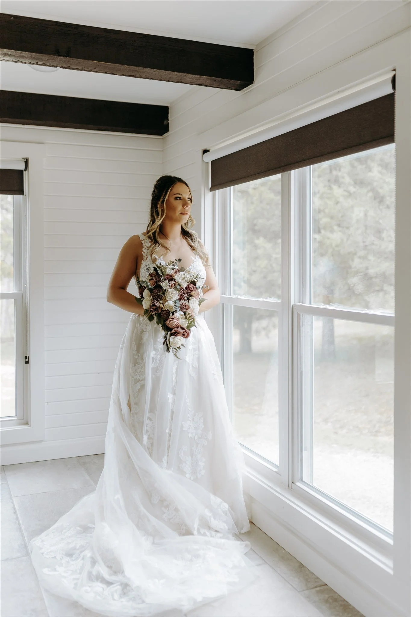 Portrait of bride standing by a large window in a white room, holding a bouquet of flowers, wearing a wedding dress with lace details at Rolla, Missouri rustic wedding venue, The Village. Photos by Columbia, MO Photographer Liv Strange Photography.