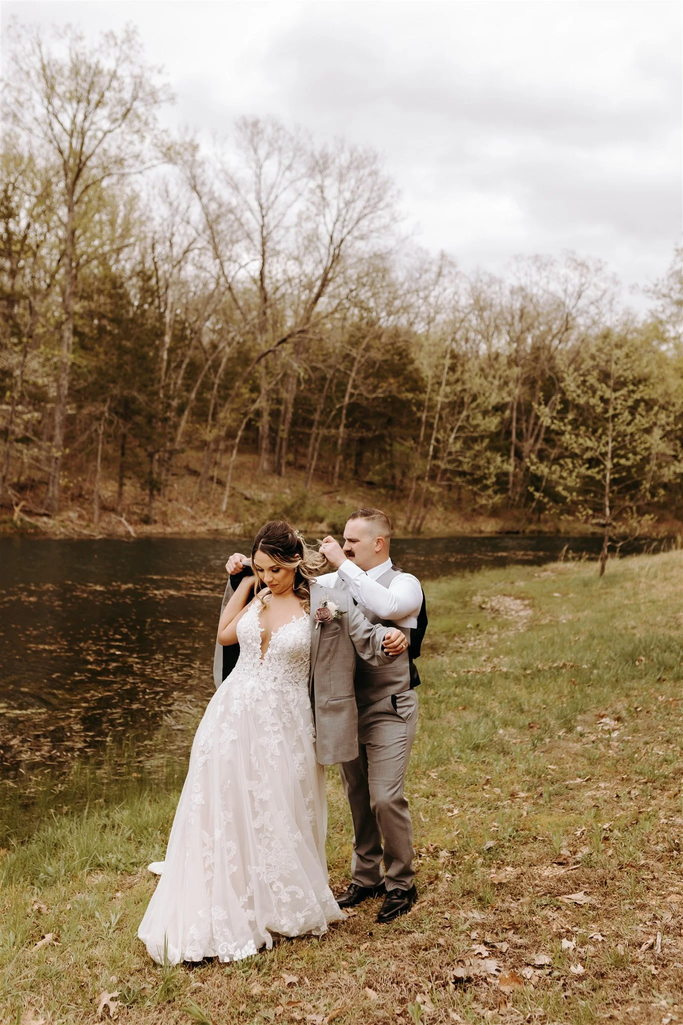 A bride and groom standing outdoors near a river, with trees in the background. The groom is helping the bride adjust her hair at Rolla, MO rustic wedding venue, The Village. Photos by Columbia, MO Photographer Liv Strange Photography.