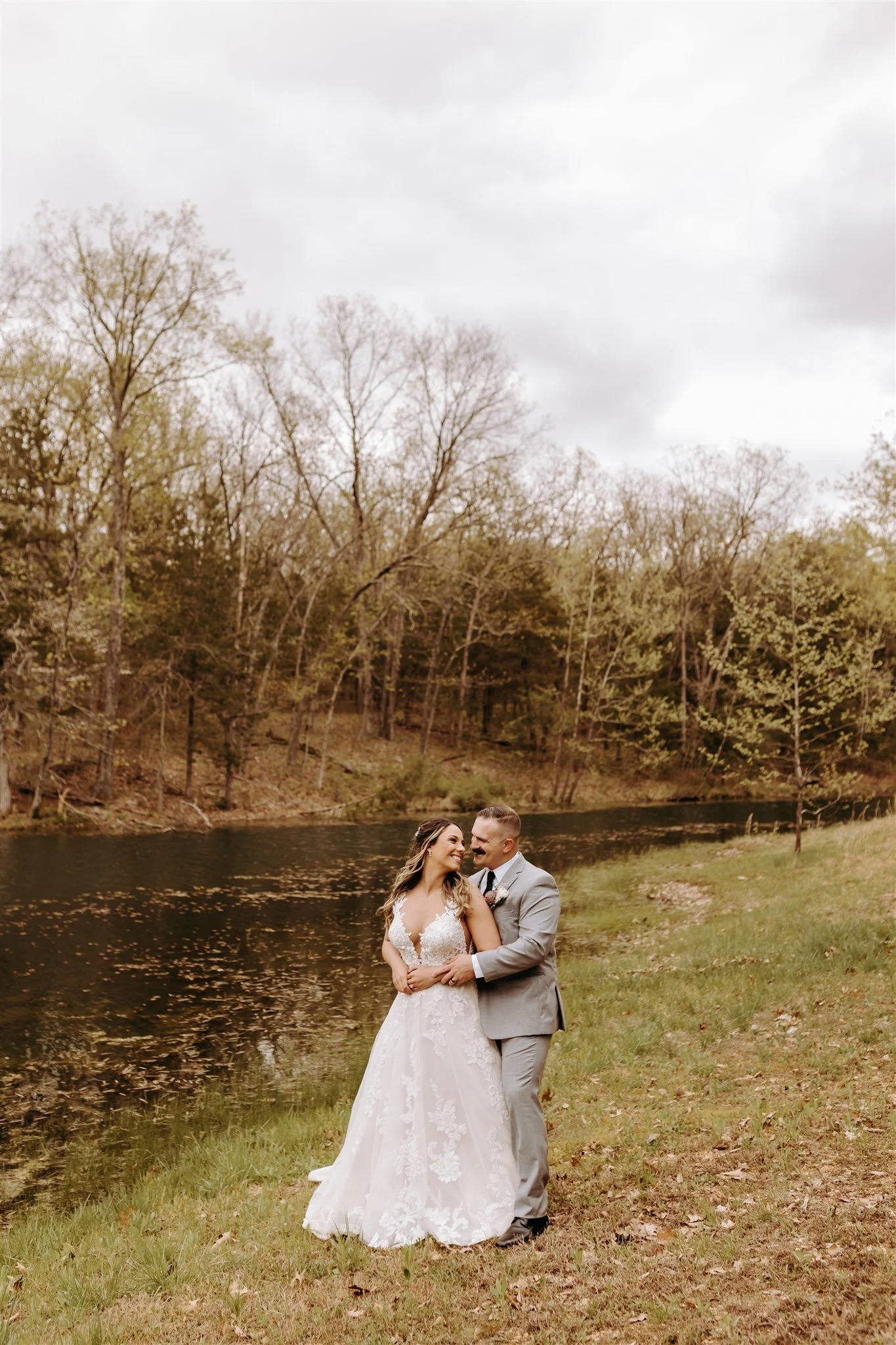 A bride and groom standing on grass by a lake, smiling and embracing outdoors surrounded by trees with fall foliage at Rolla, MO rustic wedding venue, The Village. Photos by Columbia, MO Photographer Liv Strange Photography.