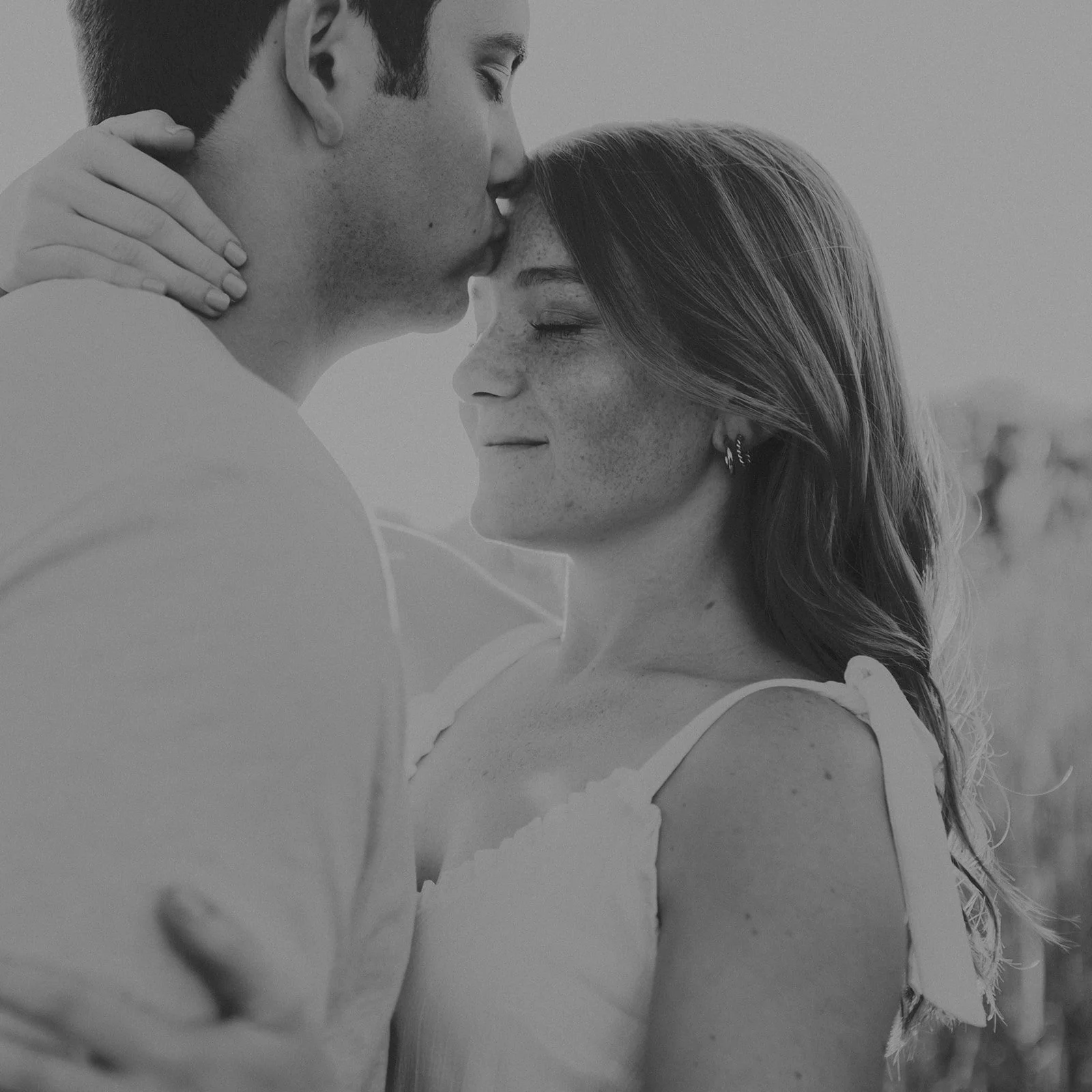 A black and white photo of a man kissing a woman on her forehead outdoors, with the woman smiling softly, eyes closed, wearing a sleeveless top with bow details on the straps during their engagement photography session at Rock Bridge State Park.