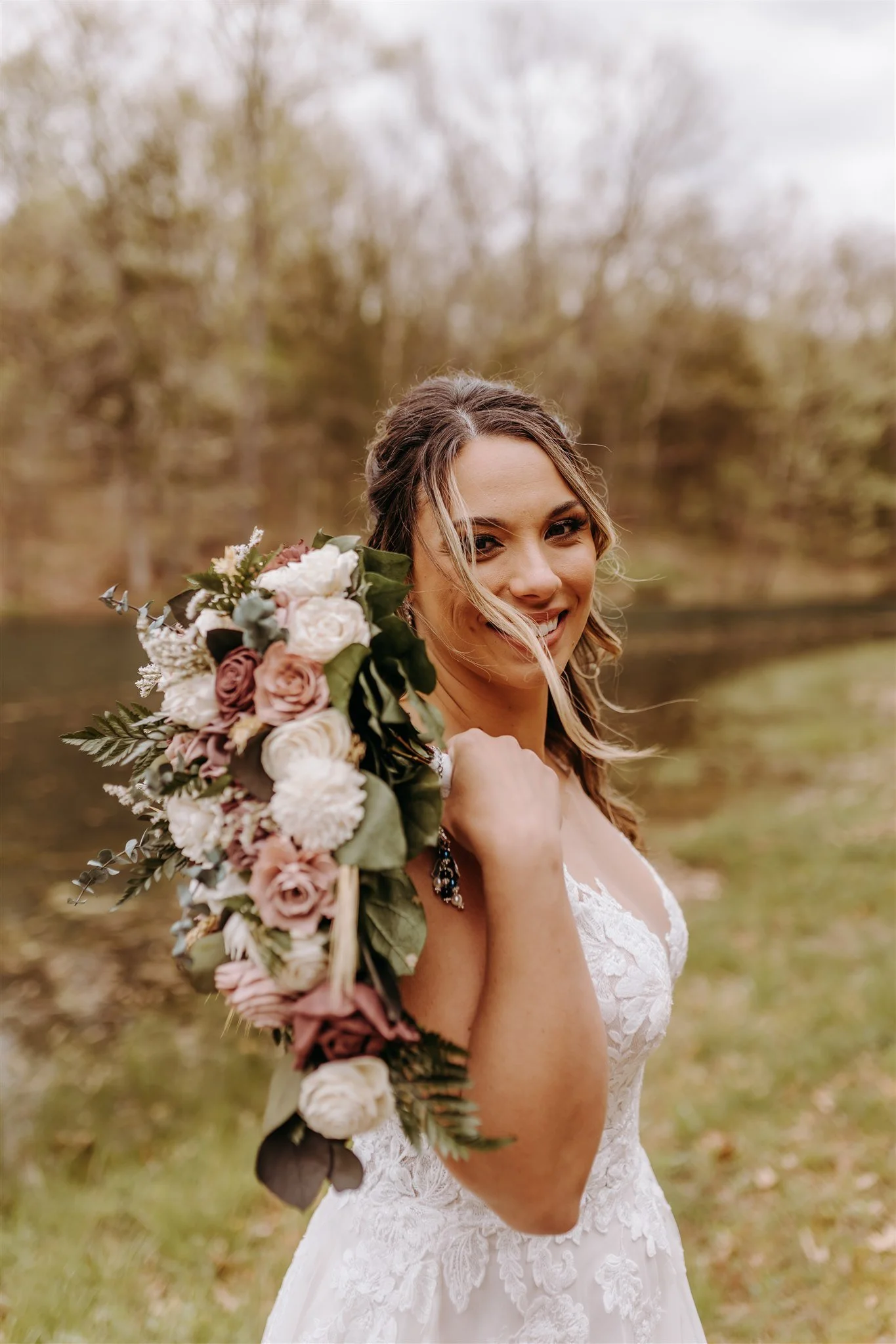 A bride outdoors, holding a large bouquet of flowers on her shoulder, smiling at the camera, with trees and a lake in the background at Rolla, MO rustic wedding venue, The Village. Photos by Columbia, MO Photographer Liv Strange Photography.