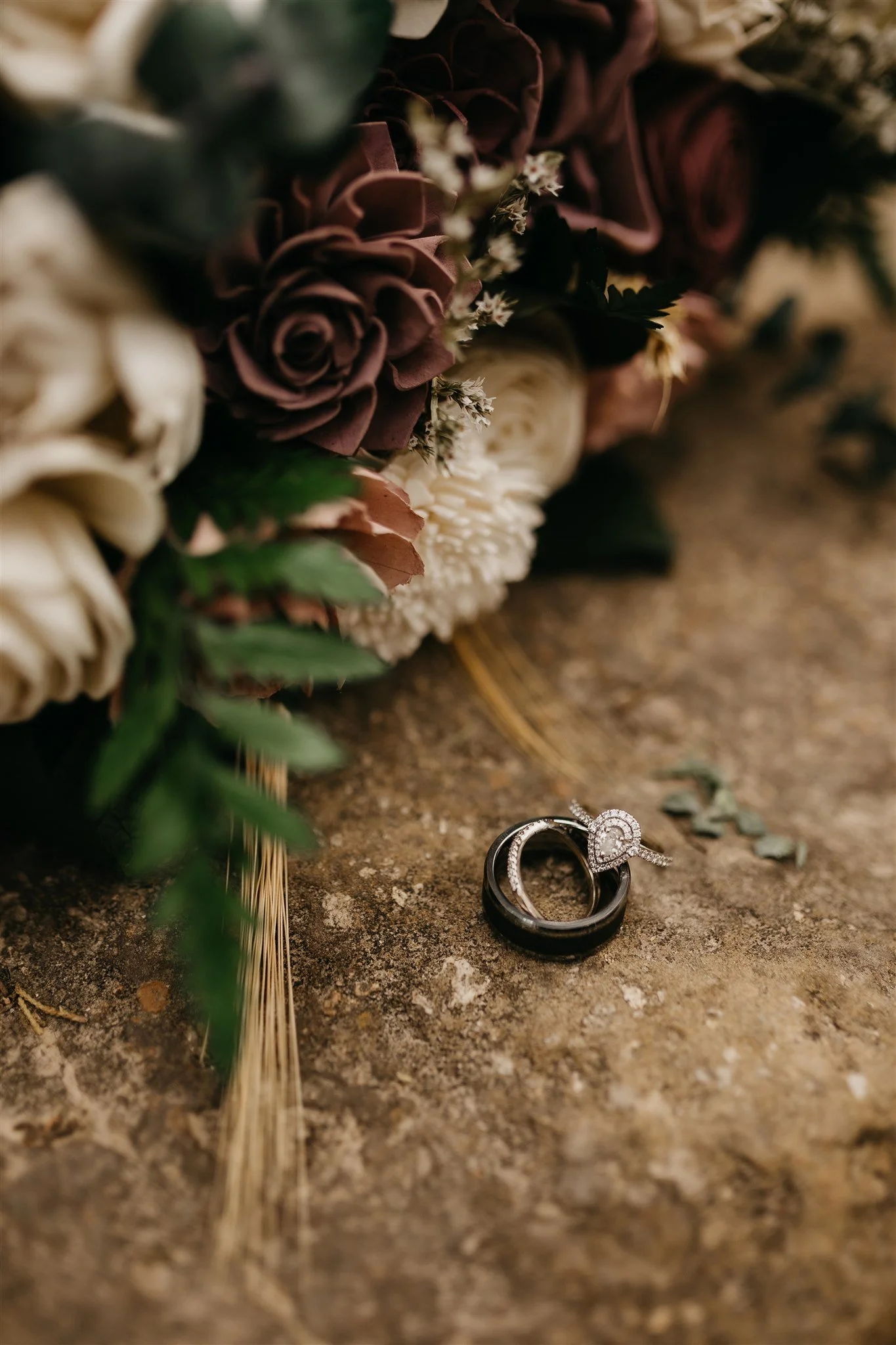 Close-up of wedding rings and engagement ring on a stone surface with a blurred bouquet of flowers in the background at Rolla, Missouri rustic wedding venue, The Village. Photos by Columbia, MO Photographer Liv Strange Photography.