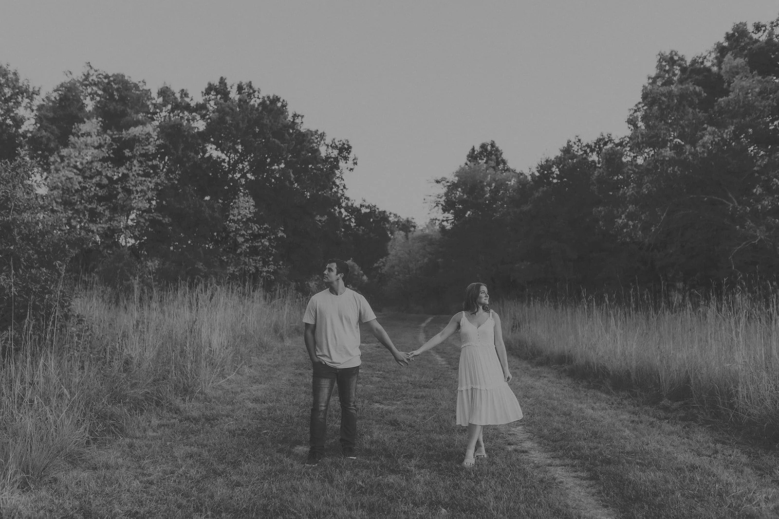 A black-and-white photo of a man and woman holding hands while walking along a grassy trail surrounded by tall grass and trees during an engagement photography session at Rock Bridge State Park in Columbia, MO.