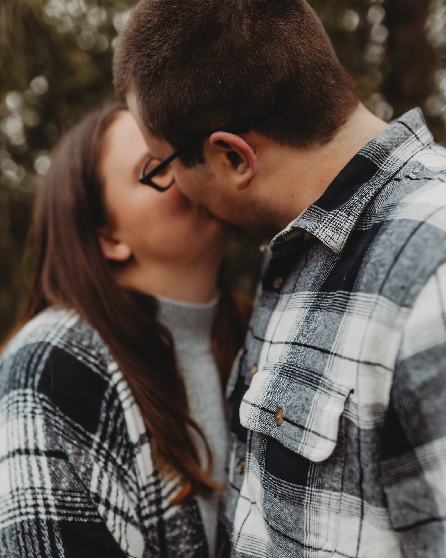 A couple is sharing a kiss outdoors with the man wearing glasses and a plaid shirt, and the woman wearing a gray sweater and a plaid shirt at their engagement photography session at Queeny Park in STL.
