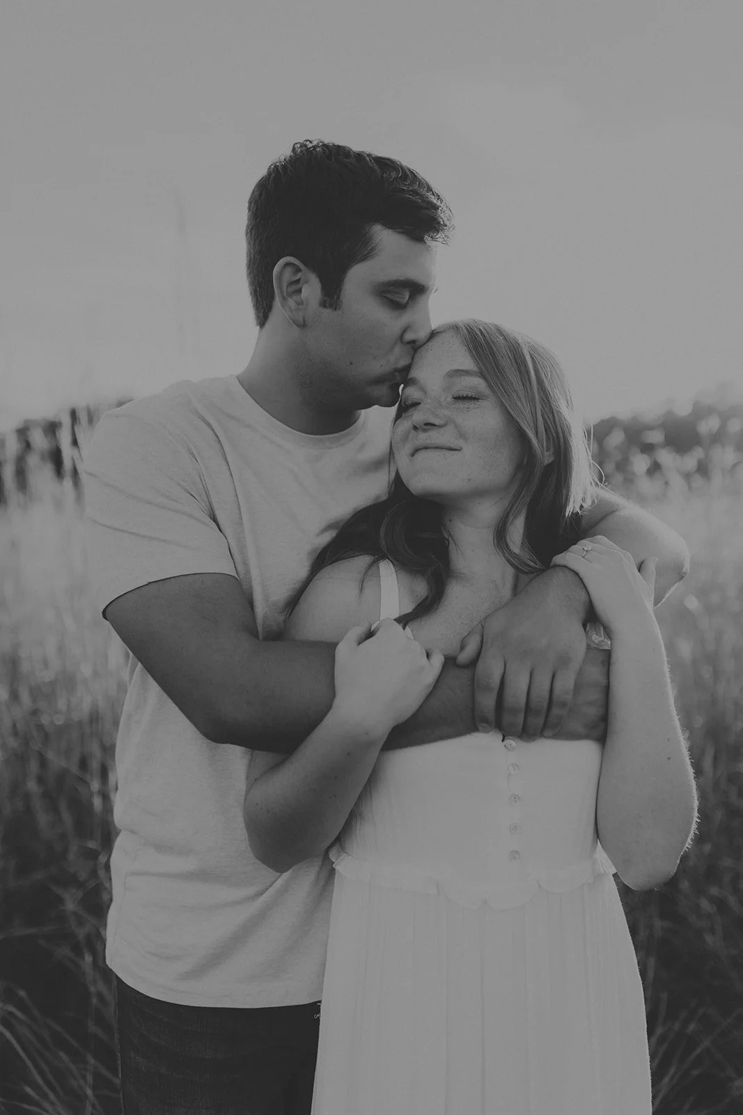 A black-and-white photo of a man gently kissing a woman on her forehead while they embrace outdoors during sunset, with grassy field in the background during their engagement photography session at Rock Bridge State Park in Columbia, MO.