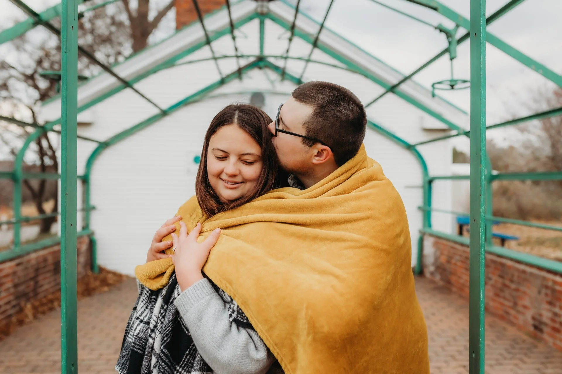 A man and woman hugging under a yellow blanket on a brick pathway, with a green metal arch overhead and trees in the background at their engagement photography session at Queeny Park in STL.