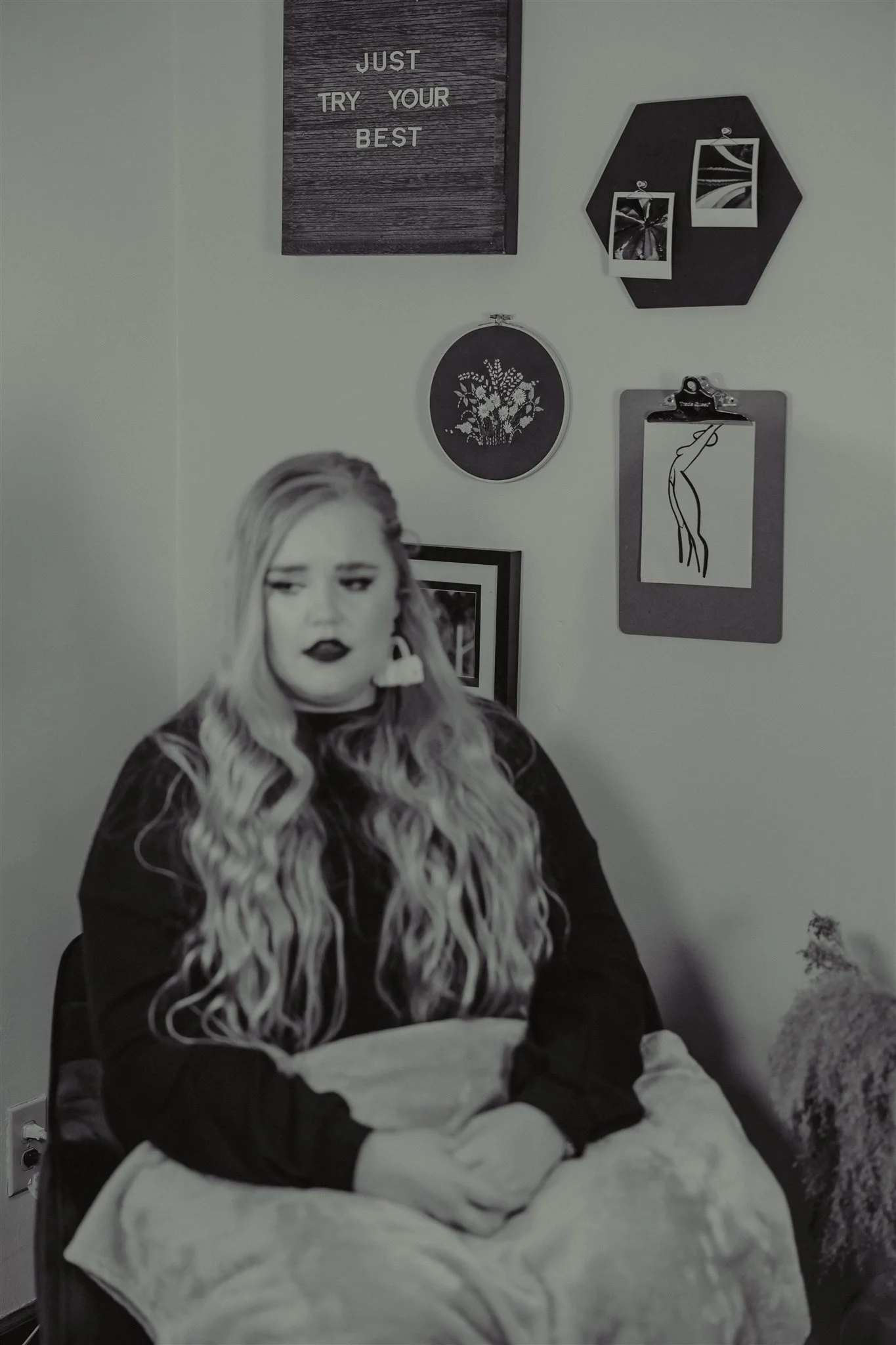 A black and white photograph of a woman with wavy long hair, sitting with her hands folded in her lap, in front of a decorative wall with framed pictures and wall decor during a trauma informed photography session in Columbia, Missouri.