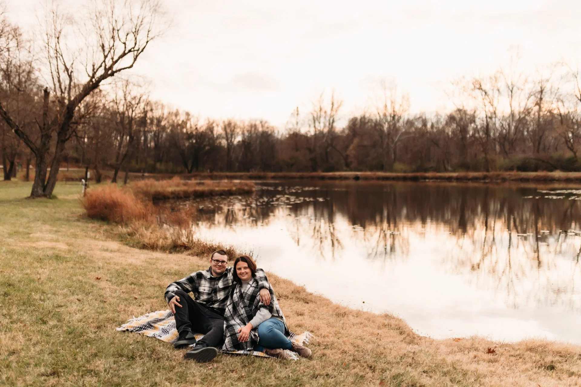A couple sitting on a blanket by a lake during autumn with bare trees and a softly lit sky at their engagement photography session at Queeny Park in STL.