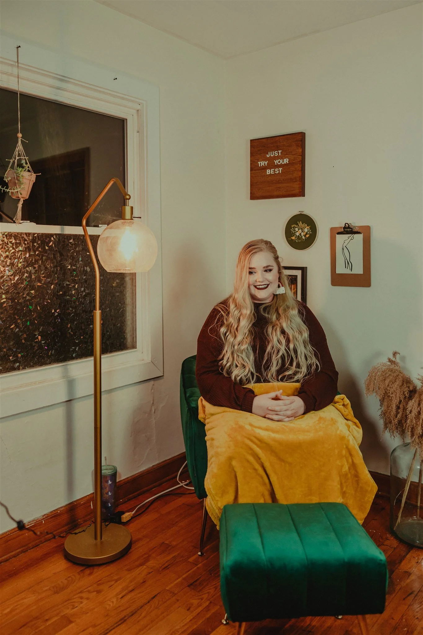 A young woman with long, wavy blonde hair and dark lipstick sits on a chair covered with a yellow blanket, smiling. She is in a cozy room during a trauma informed photography session in Columbia, Missouri.