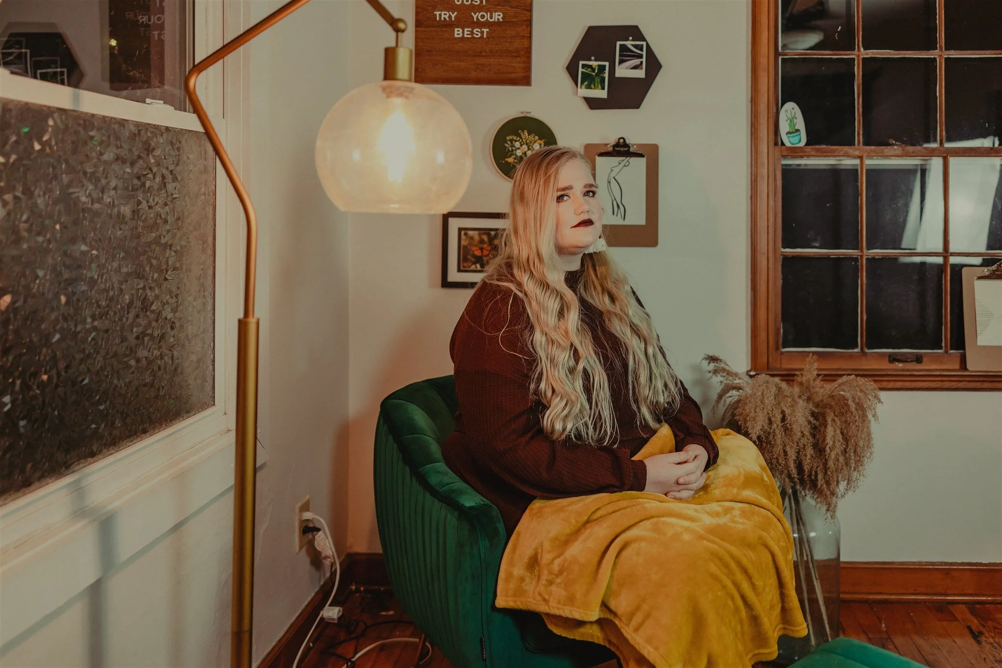 A woman with long wavy blonde hair and dark makeup, sitting on a green upholstered chair with her hands clasped, covered with a yellow blanket, in a cozy room during a trauma informed photography session in Columbia, MO.
