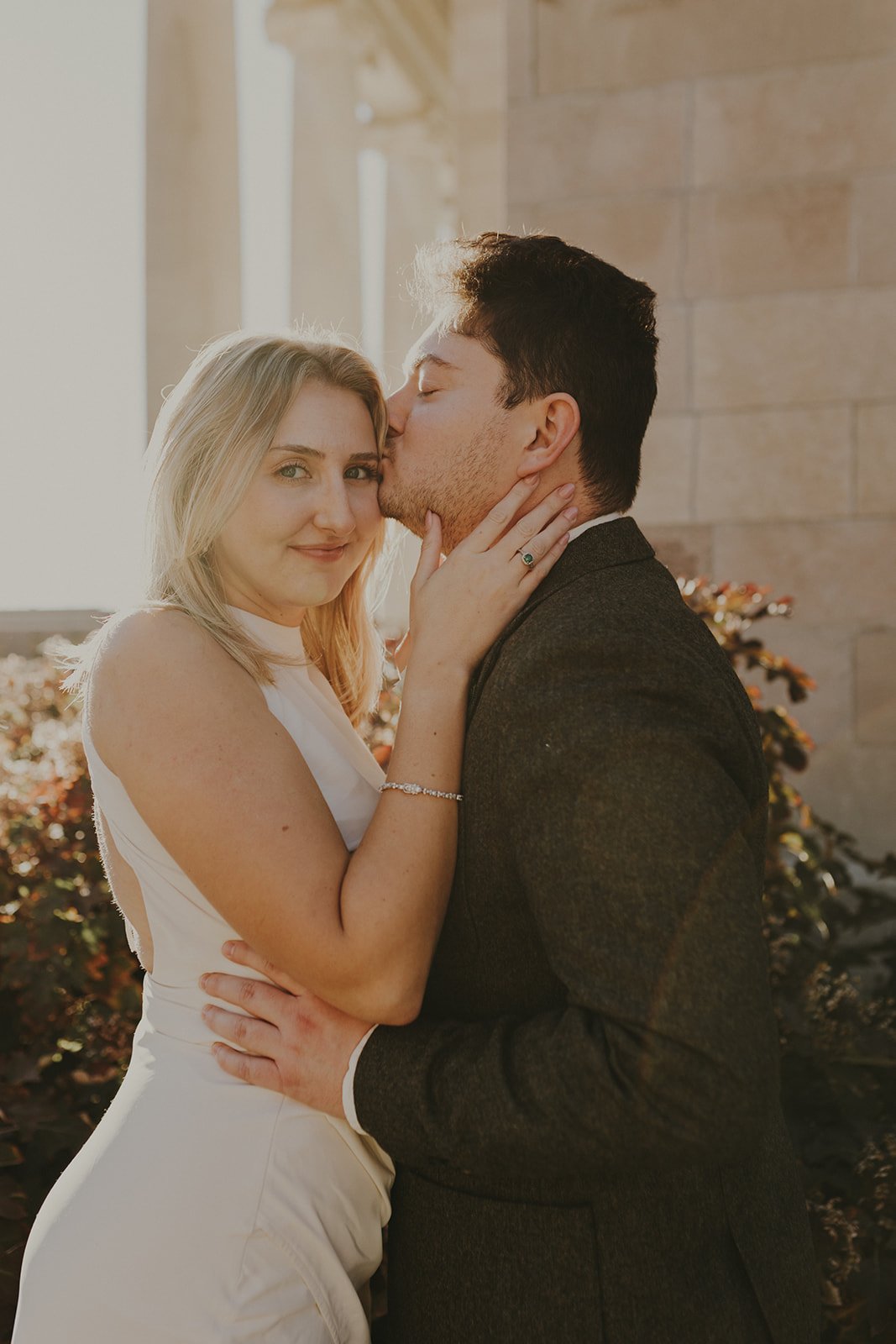 A couple embracing, with the man kissing the woman on the forehead and the woman smiling at the camera, outdoors under sunlight.