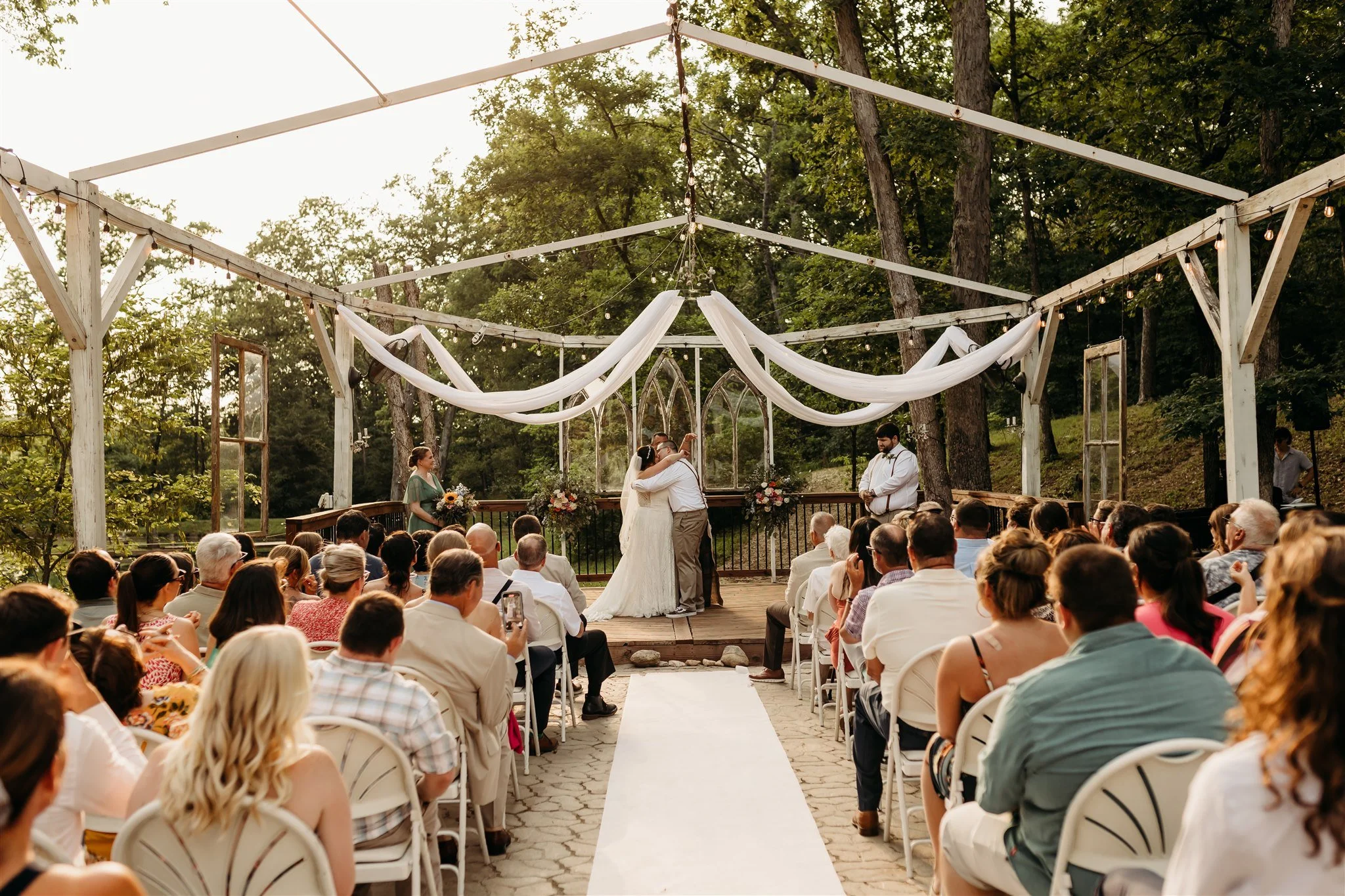 A wedding ceremony outdoors on a wooden platform, with the bride and groom embracing, surrounded by guests seated in white chairs, trees in the background, and decorative white drapes overhead at Foristell, MO wedding venue, Renaissance Wine Garden.