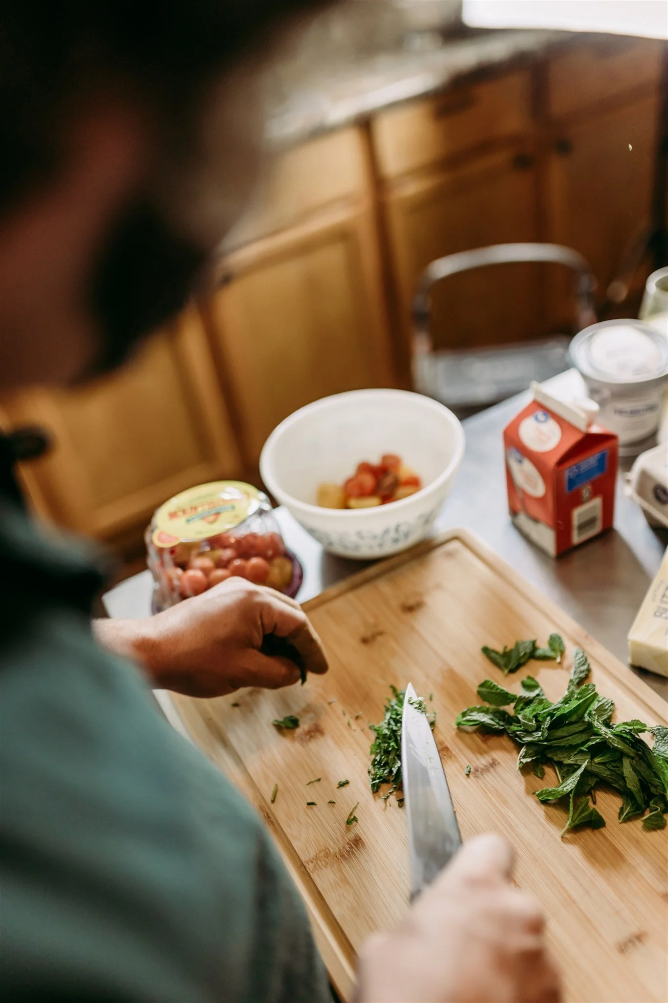 A person chopping fresh herbs on a wooden cutting board in a kitchen with various dairy products and fruit in the background.