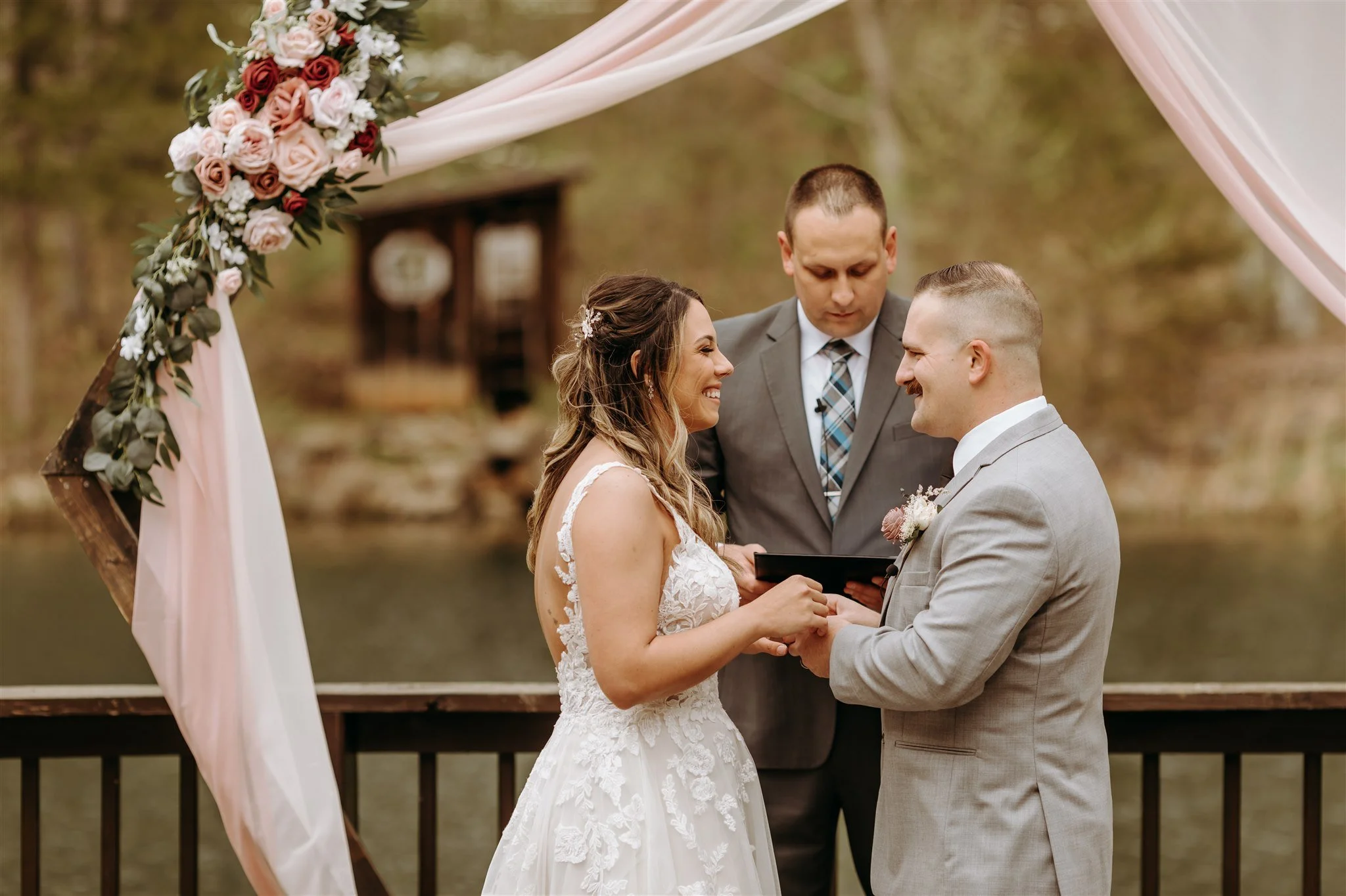 A bride and groom exchanging vows during their outdoor wedding ceremony by a lake with trees in the background at Rolla, Missouri rustic wedding venue, The Village. Photos by Columbia, MO Photographer Liv Strange Photography.