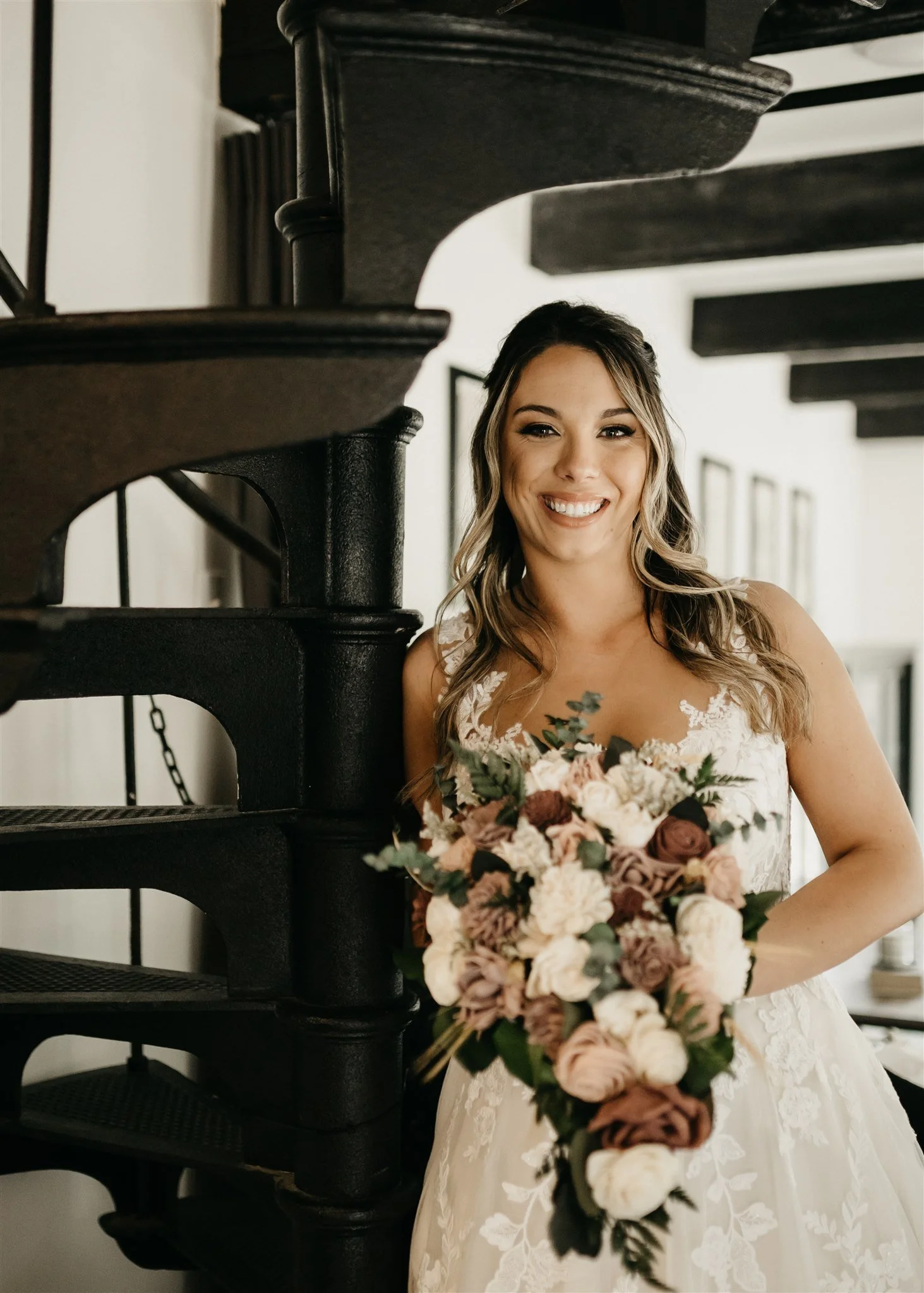 A smiling bride with wavy hair, wearing a white lace wedding dress, holding a bouquet, standing beside a black staircase railing at Rolla, Missouri rustic wedding venue, The Village. Photos by Columbia, MO Photographer Liv Strange Photography.