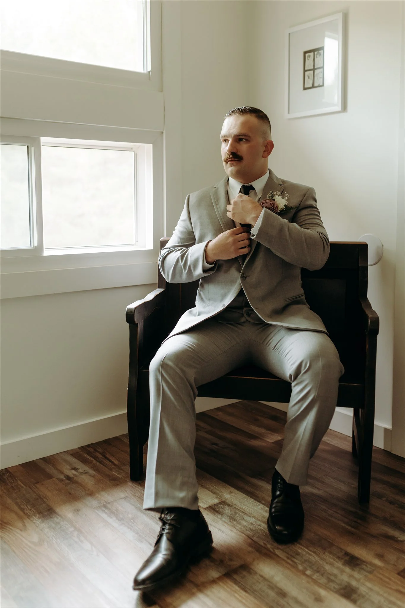 Portrait of groom in a light gray suit sitting on a dark wooden chair near a window, adjusting his tie at Rolla, Missouri rustic wedding venue, The Village. Photos by Columbia, MO Photographer Liv Strange Photography.