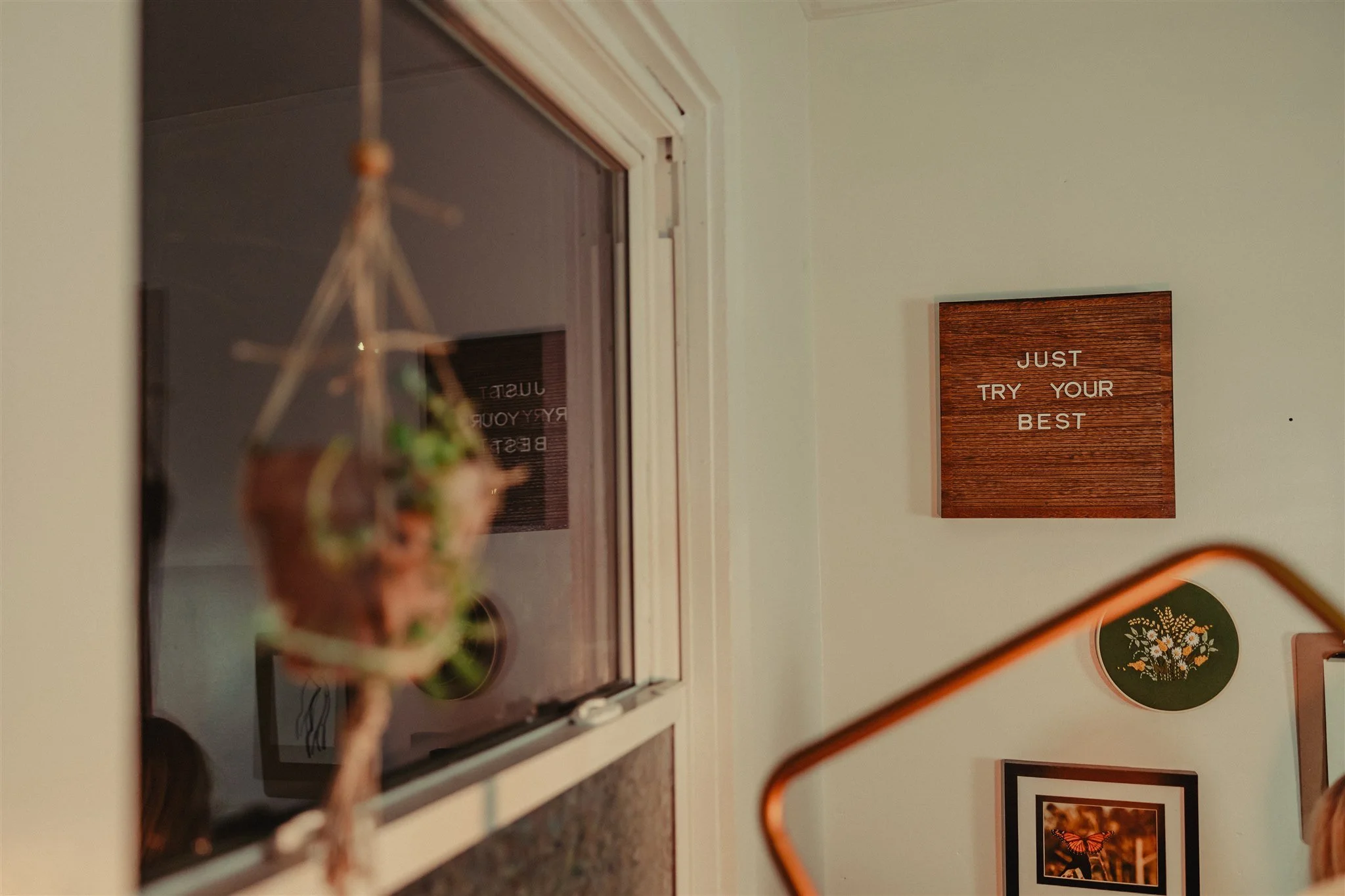 Interior wall with motivational signs, including a wooden square sign that says 'JUST TRY YOUR BEST,' and picture frames with art and photographs, a window with a hanging plant outside during a trauma informed photography session in Columbia, MO