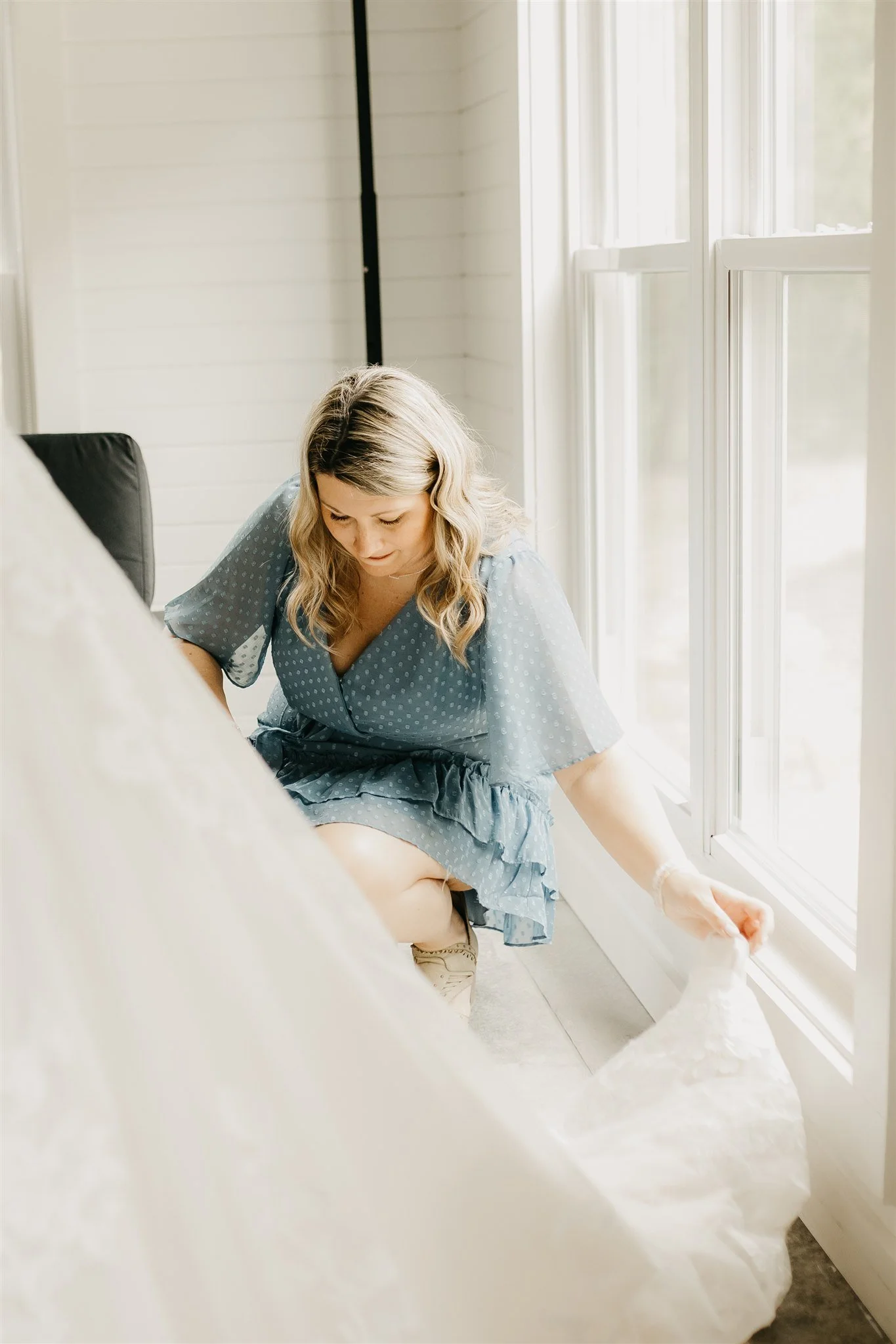Woman in a blue polka-dot dress sitting on the floor near a window, folding or arranging white fabric or a sheet at Rolla, Missouri rustic wedding venue, The Village. Photos by Columbia, MO Photographer Liv Strange Photography.