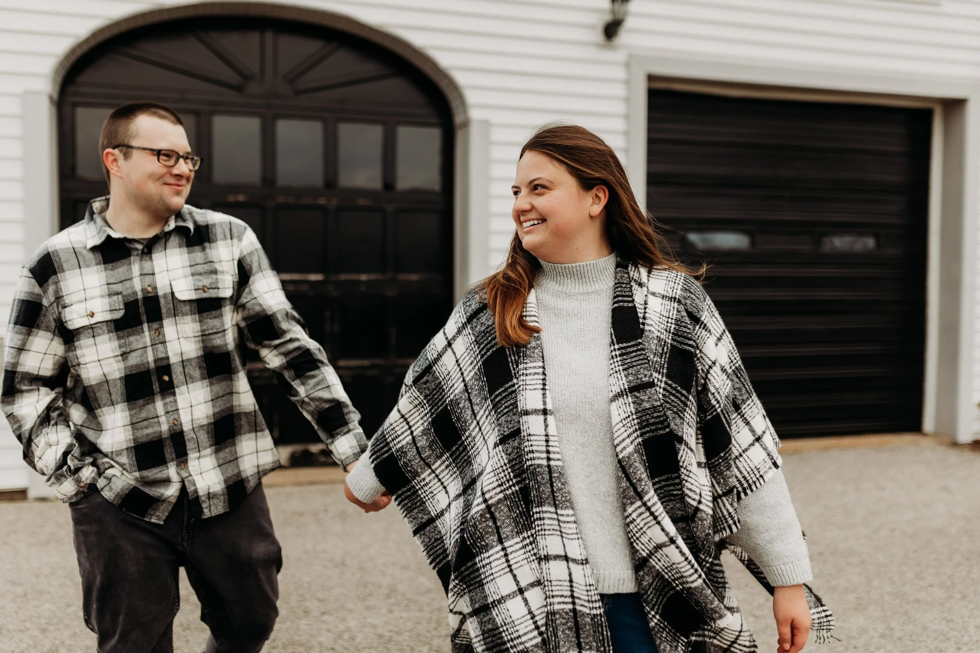 A smiling couple holding hands in front of a garage, wearing plaid shirts and casual clothing at their engagement photography session at Queeny Park in STL.