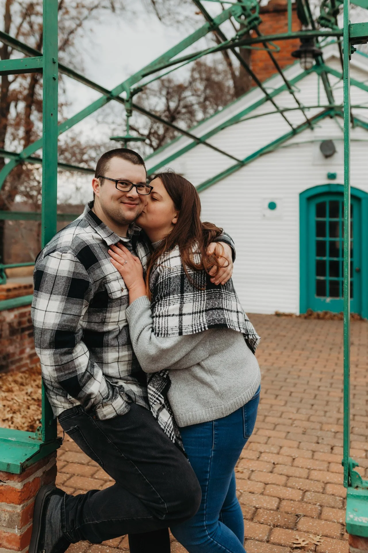 A couple dressed in plaid and grey sweaters sharing a kiss outdoors in front of a white building with blue accents, surrounded by an outdoor structure at their engagement photography session at Queeny Park in STL..