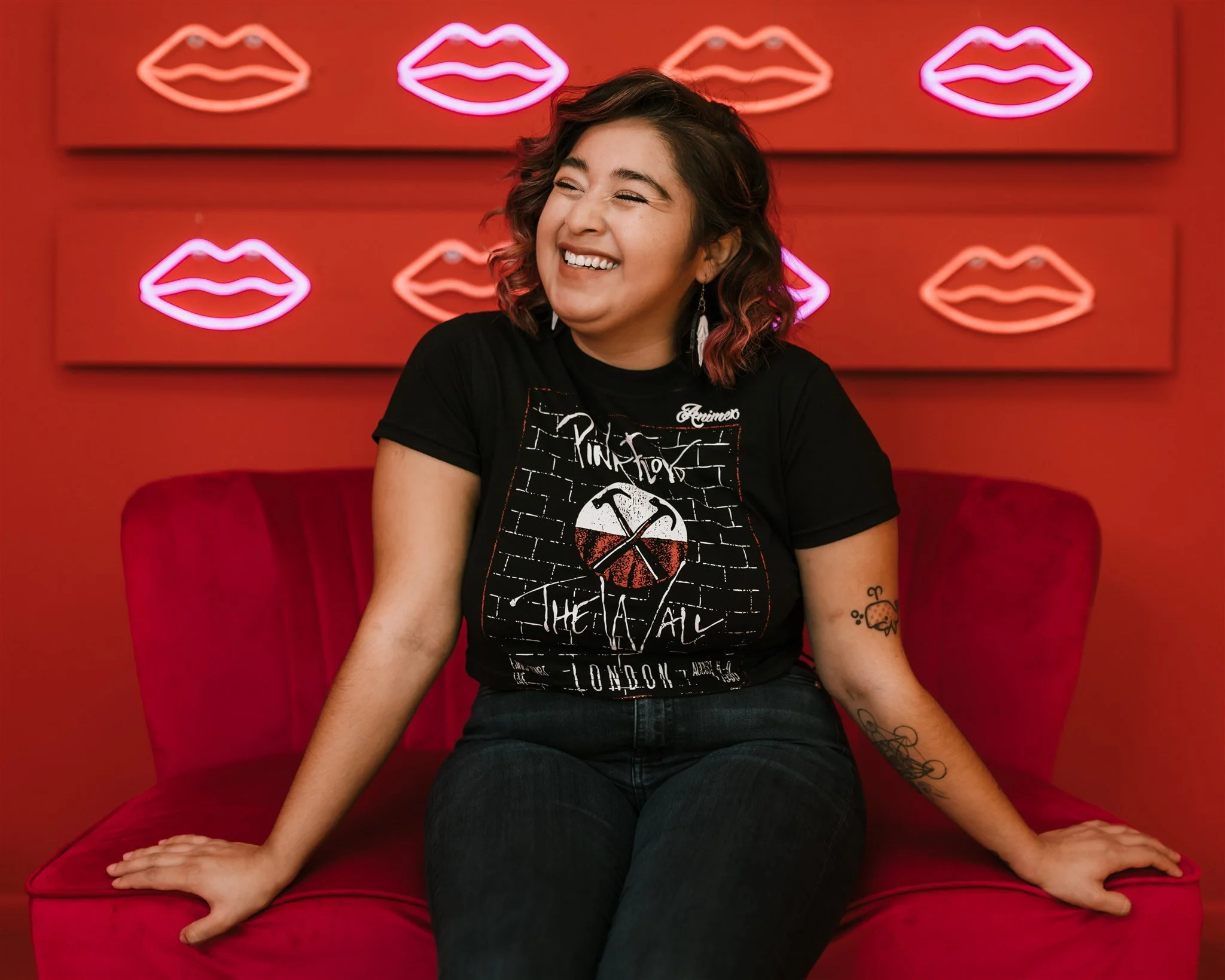 A woman with curly hair sitting on a red couch, smiling and winking with closed eyes, against a red wall decorated with neon pink lips.