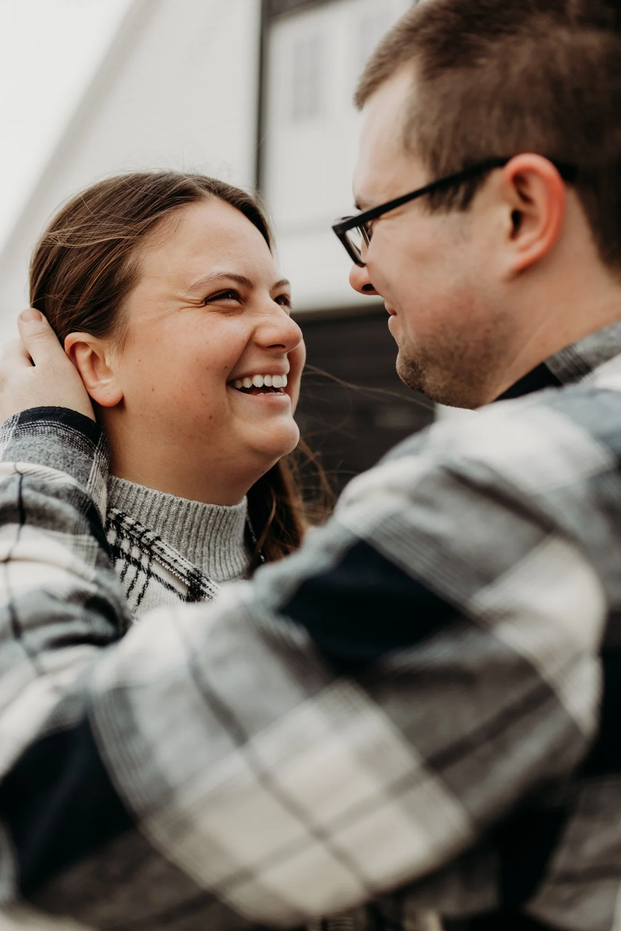 A couple smiling and looking at each other outdoors, with a building behind them at their engagement photography session at Queeny Park in STL.