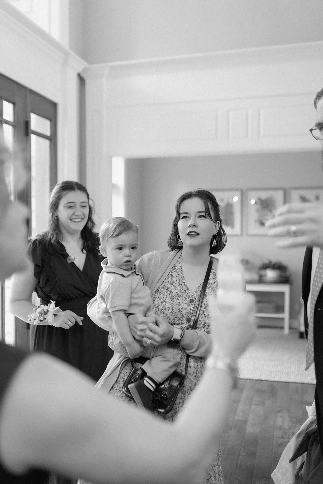 Group of people indoors, including a woman holding a young boy, with another woman behind them smiling, and a man on the right partially visible at an intimate backyard wedding in Columbia, MO.
