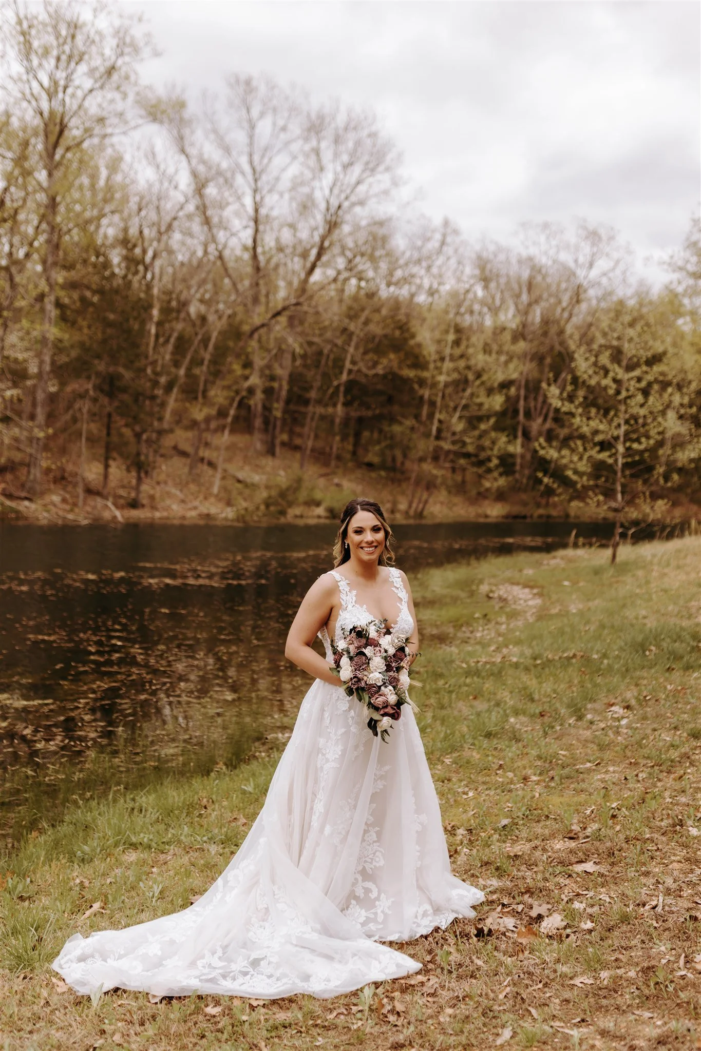 A woman in a wedding dress holding a bouquet outdoors near a river, with trees in the background at Rolla, MO rustic wedding venue, The Village. Photos by Columbia, MO Photographer Liv Strange Photography.