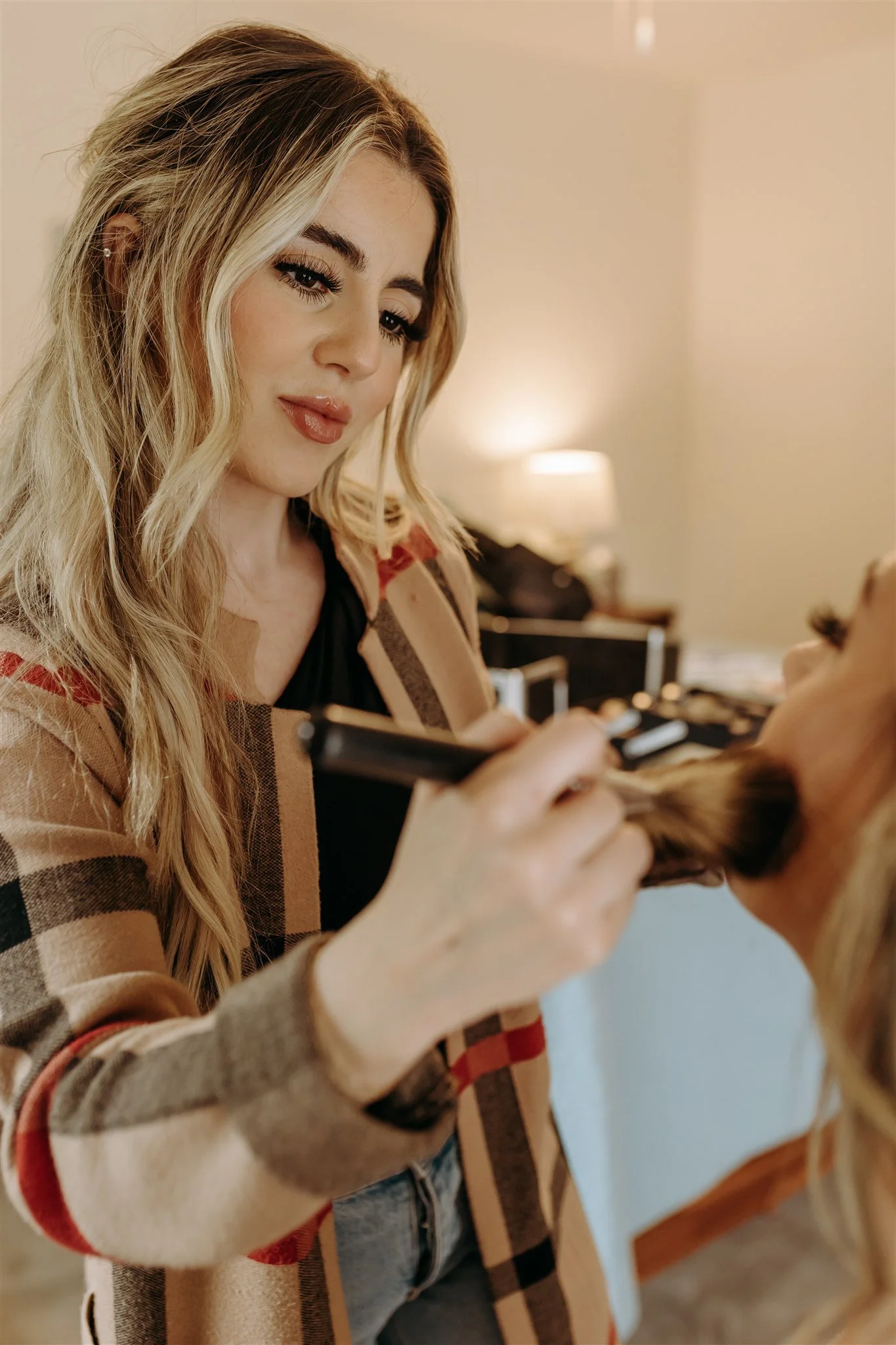 A woman with long blonde wavy hair applying makeup to another woman's face in an indoor setting at Rolla, Missouri rustic wedding venue, The Village. Photos by Columbia, MO Photographer Liv Strange Photography.