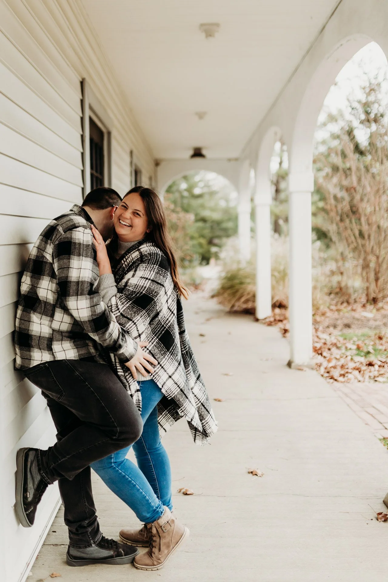 A smiling woman and a man in plaid shirts sharing a moment of laughter on a porch with white columns and beige siding, fallen leaves on the ground at their engagement photography session at Queeny Park in STL.